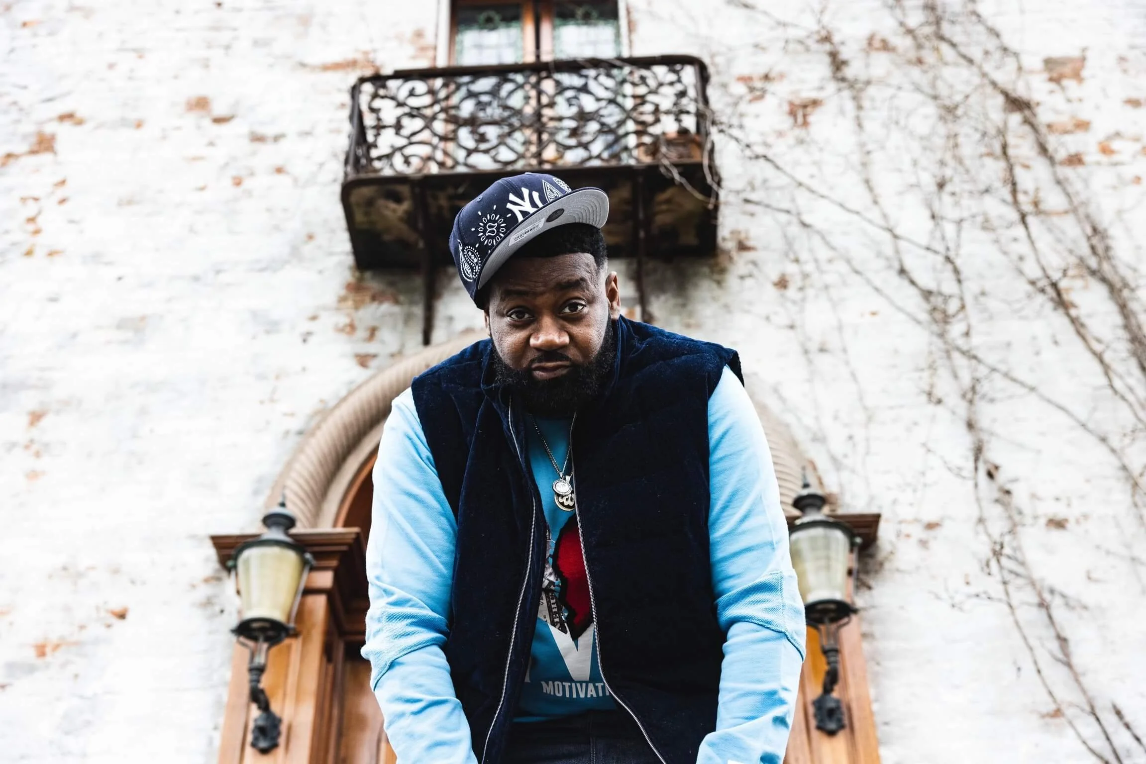A man wearing a navy New York Yankees cap, a blue shirt with a graphic, and a black vest, stands in front of a white brick wall with an arched wooden door and two lanterns, looking into the camera.