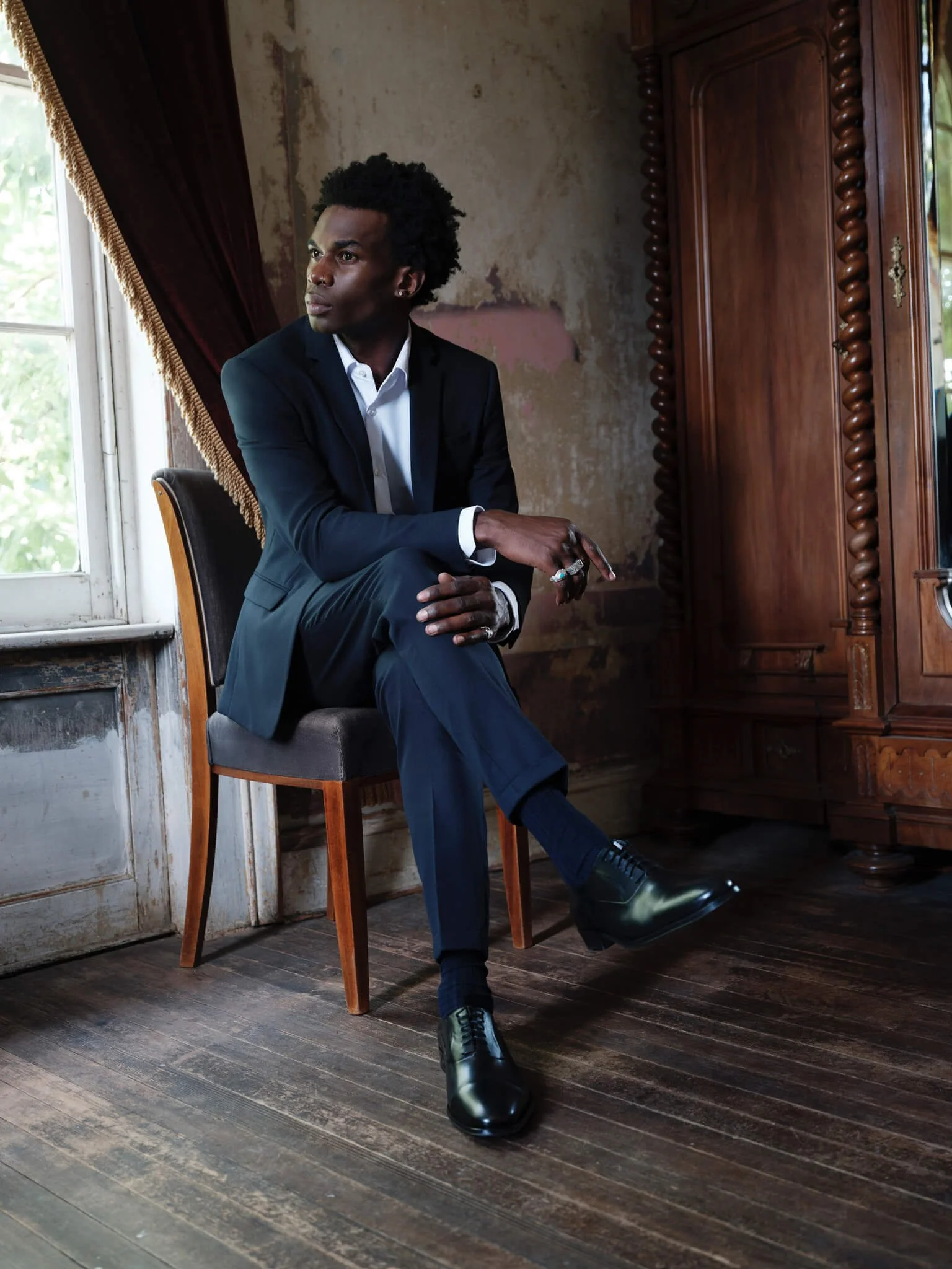 A man in a dark suit sitting on a chair in a vintage, rustic room with wooden floors and an old wardrobe, looking to the left.
