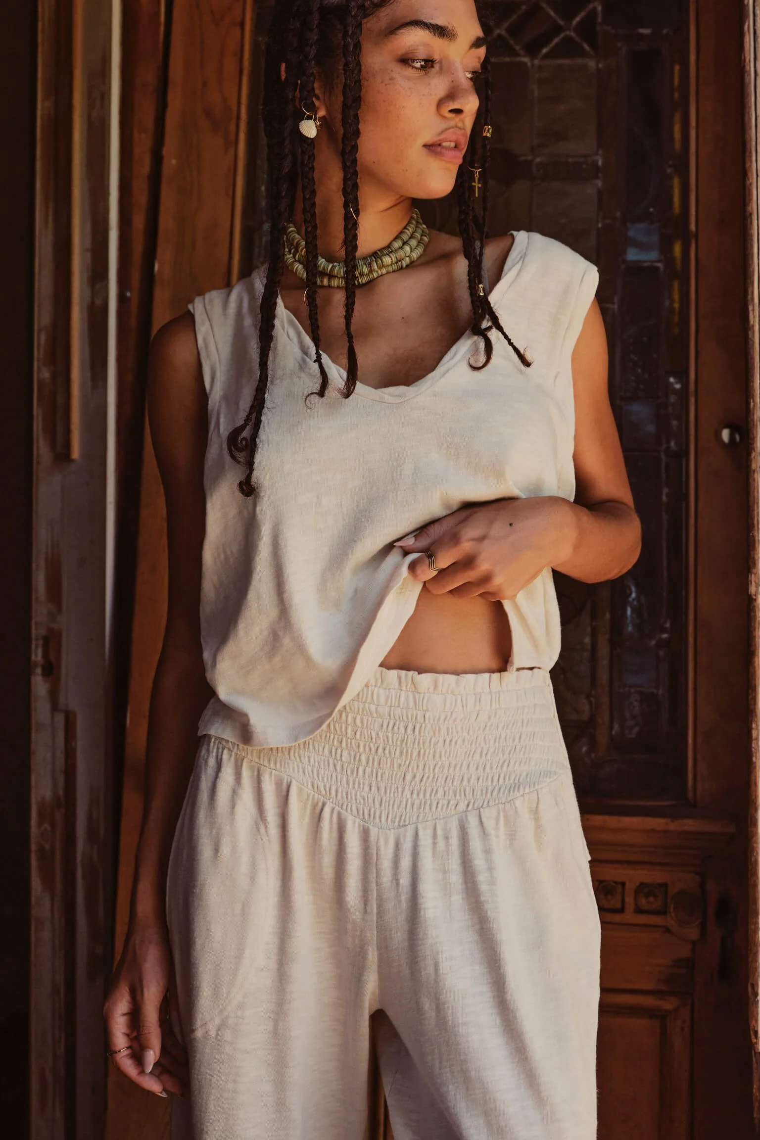 A woman with braided hair and light brown skin standing indoors near wooden doors, wearing a sleeveless cream top and loose matching cream pants, with jewelry including earrings, a choker, and rings.