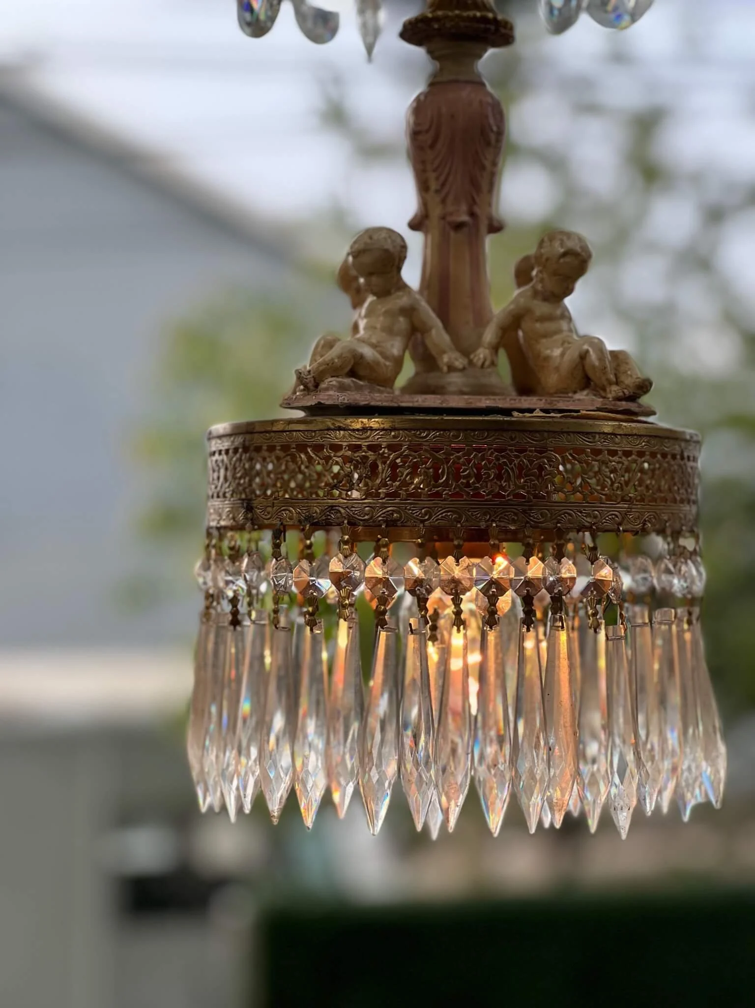 Close-up of a vintage chandelier with crystal prisms, featuring three cherub figurines on top, hanging in a room with a blurred background outside.