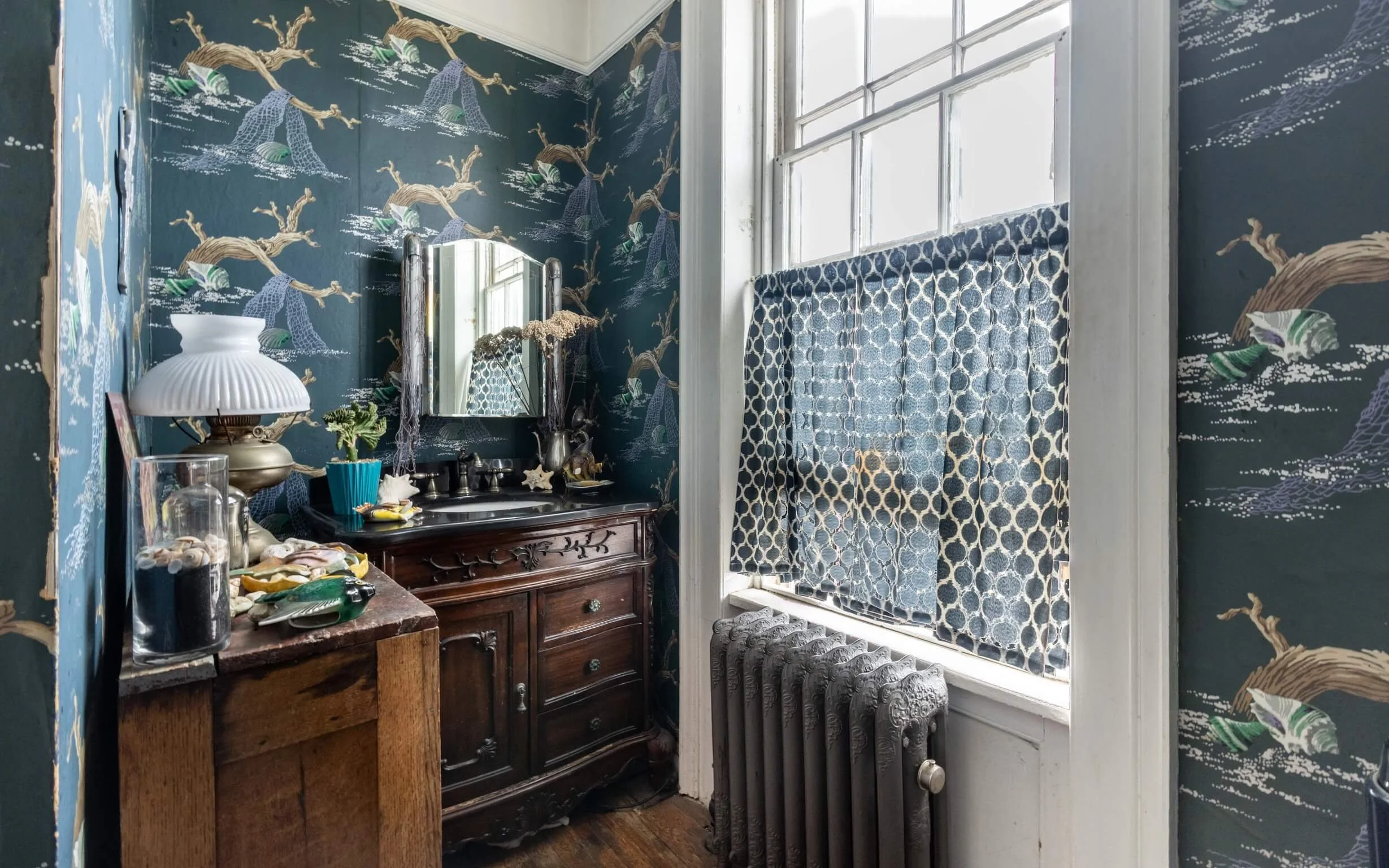 A vintage bathroom with dark wallpaper featuring a tree and ocean scene, a large window with patterned curtains, an ornate wooden vanity, a mirror, a vintage radiator, and various decorative items.