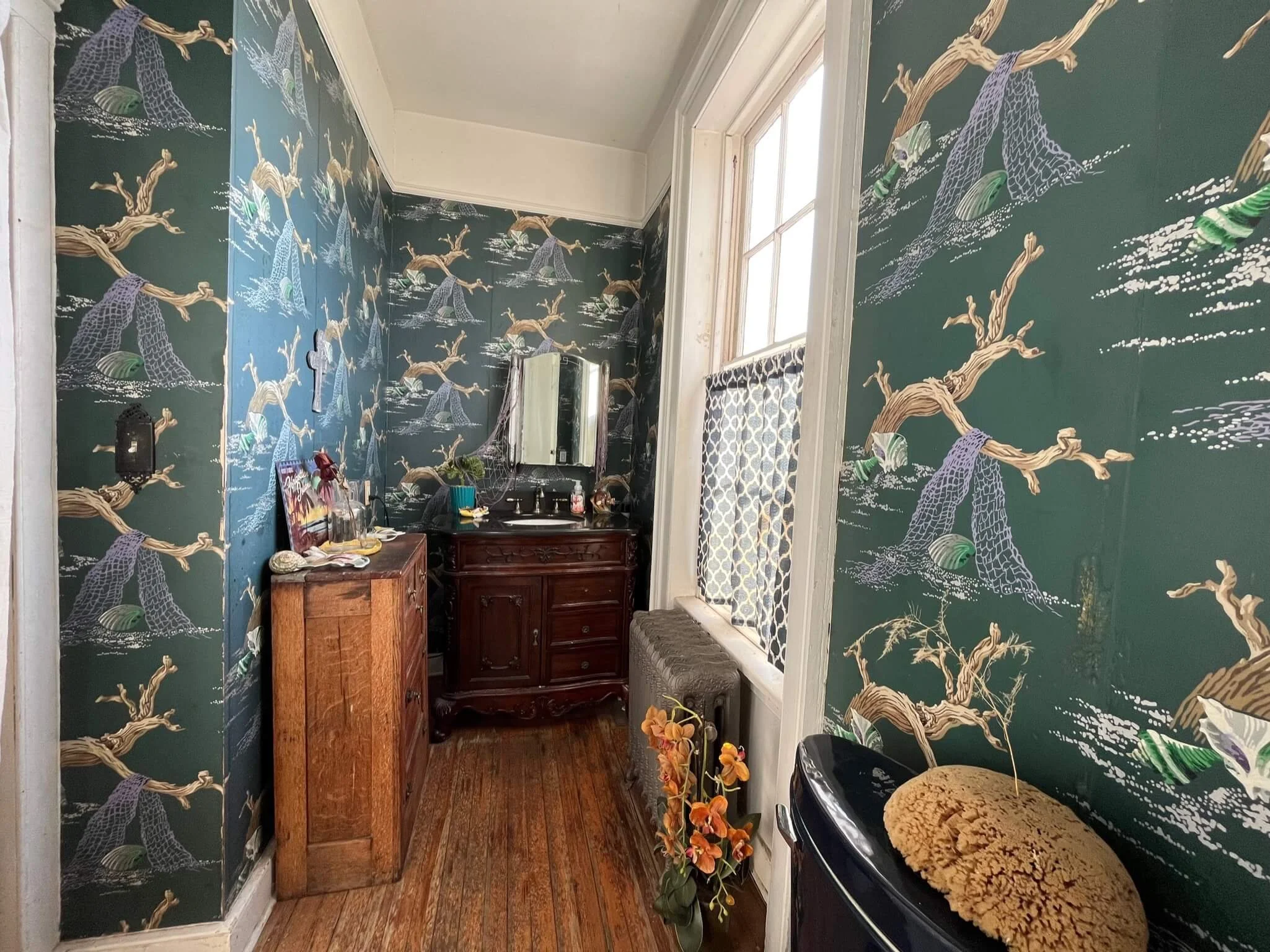 Small bathroom with dark green wallpaper decorated with trees and animals, a wooden cabinet, a mirror above a black sink, a window with patterned curtains, and various toiletries and decorations.
