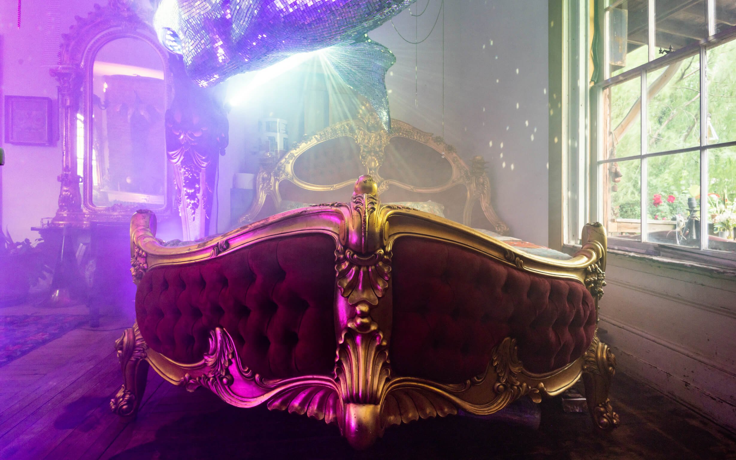 An ornate bed with a gold frame and red upholstery in a room filled with colorful light and mist. A large window on the right shows an outdoor garden with flowers.
