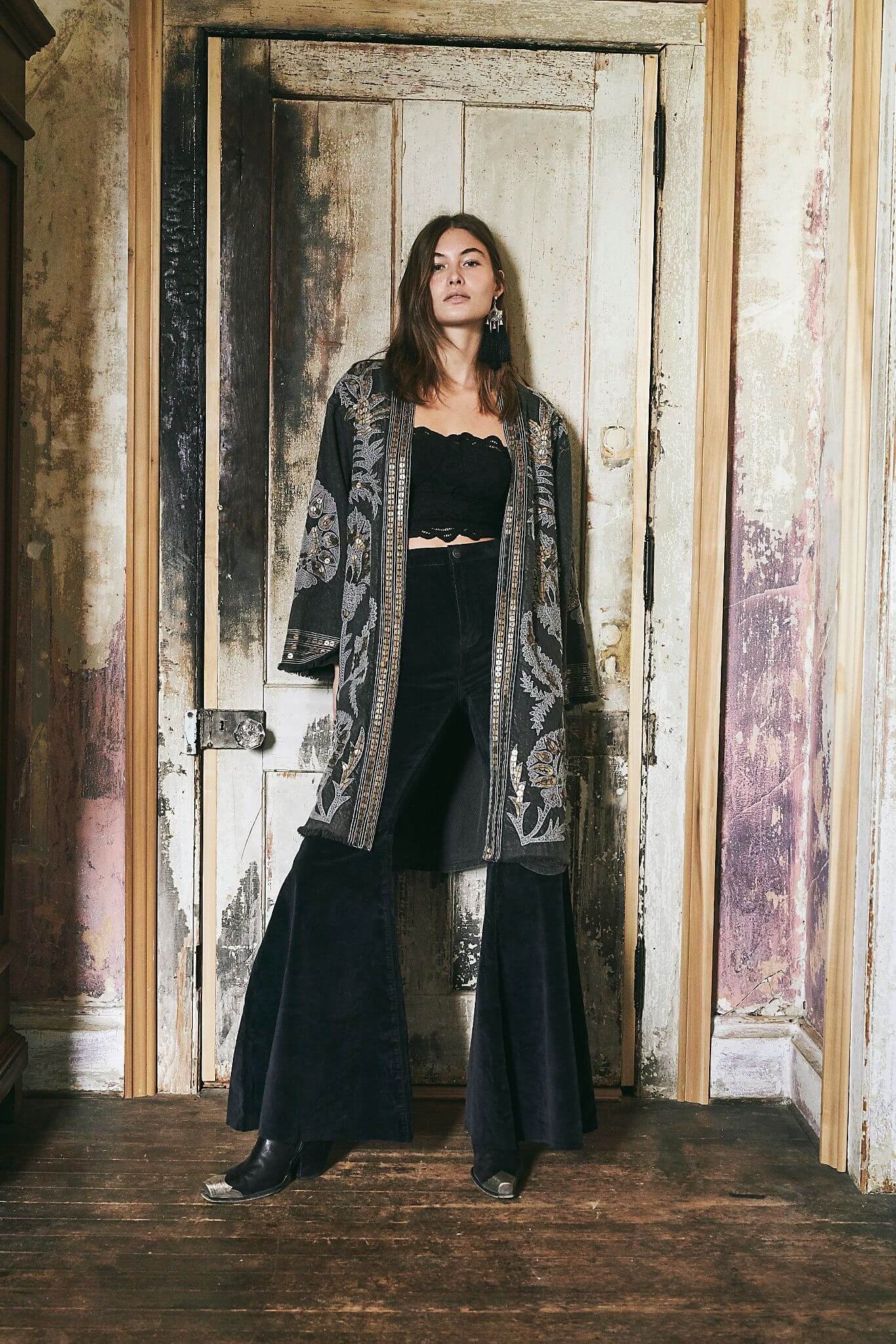 A woman standing in front of a rustic, weathered wooden door with peeling paint, wearing a black lace crop top, wide leg black pants, and a patterned long kimono, with dark boots and large earrings.