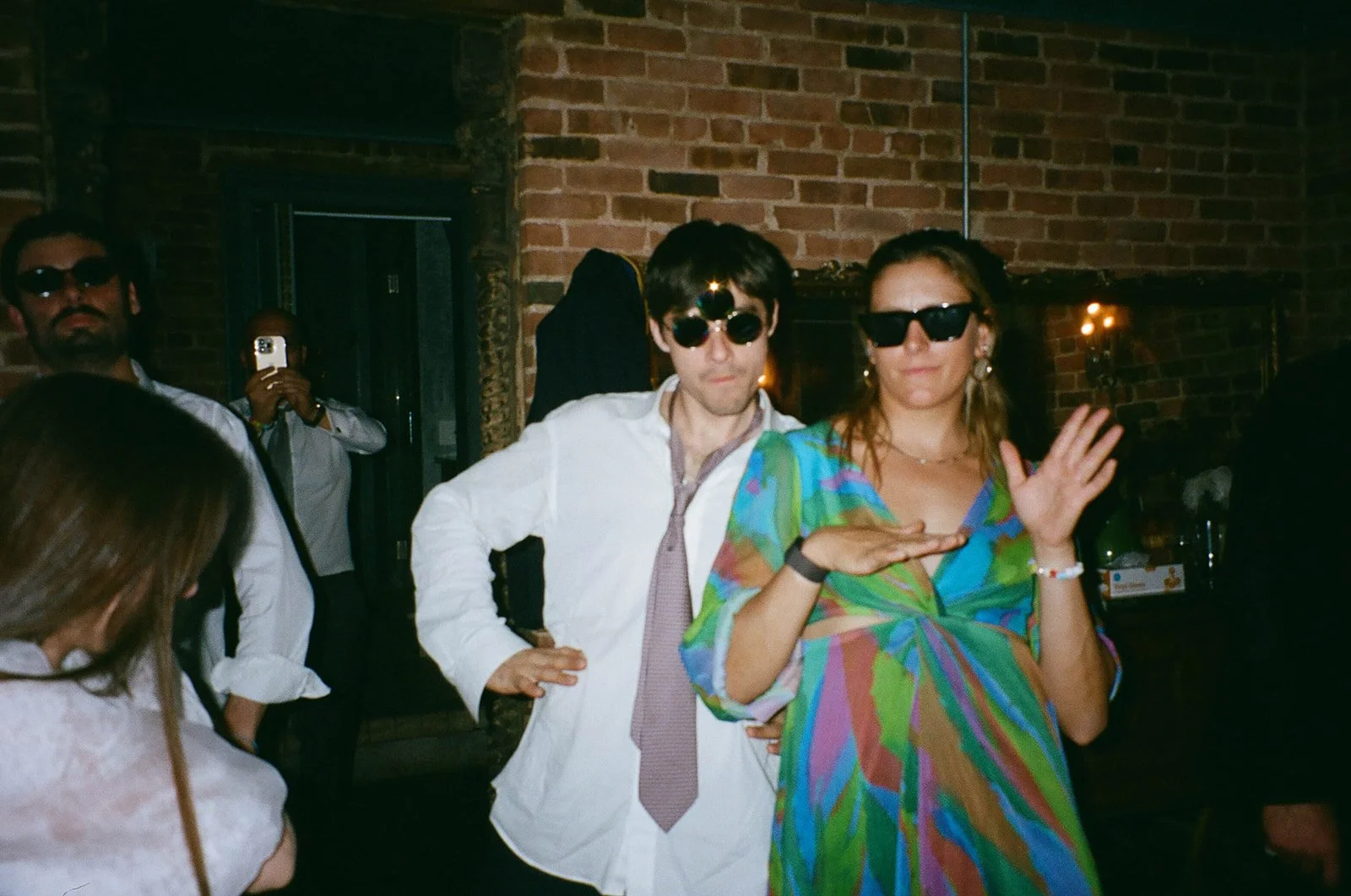 A group of people at a party or social gathering, wearing sunglasses, posing for the camera inside a dimly lit venue with brick walls.
