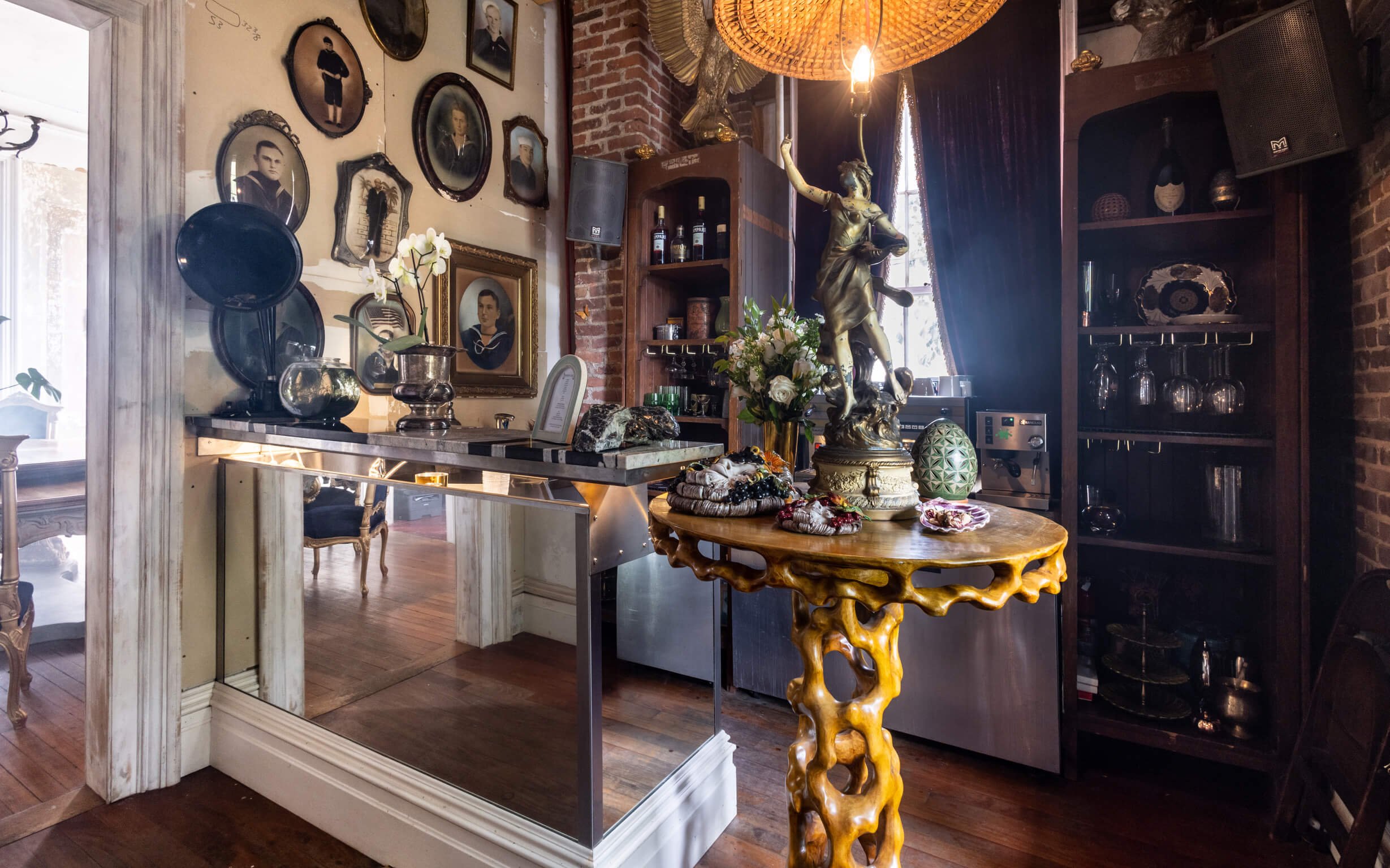 A cozy room with vintage decor, including a gallery wall of framed black-and-white photographs, a marble-topped table, an ornate wooden table with decorative items, a large sculpture of a woman holding a garland, and various glassware and bottles. Th