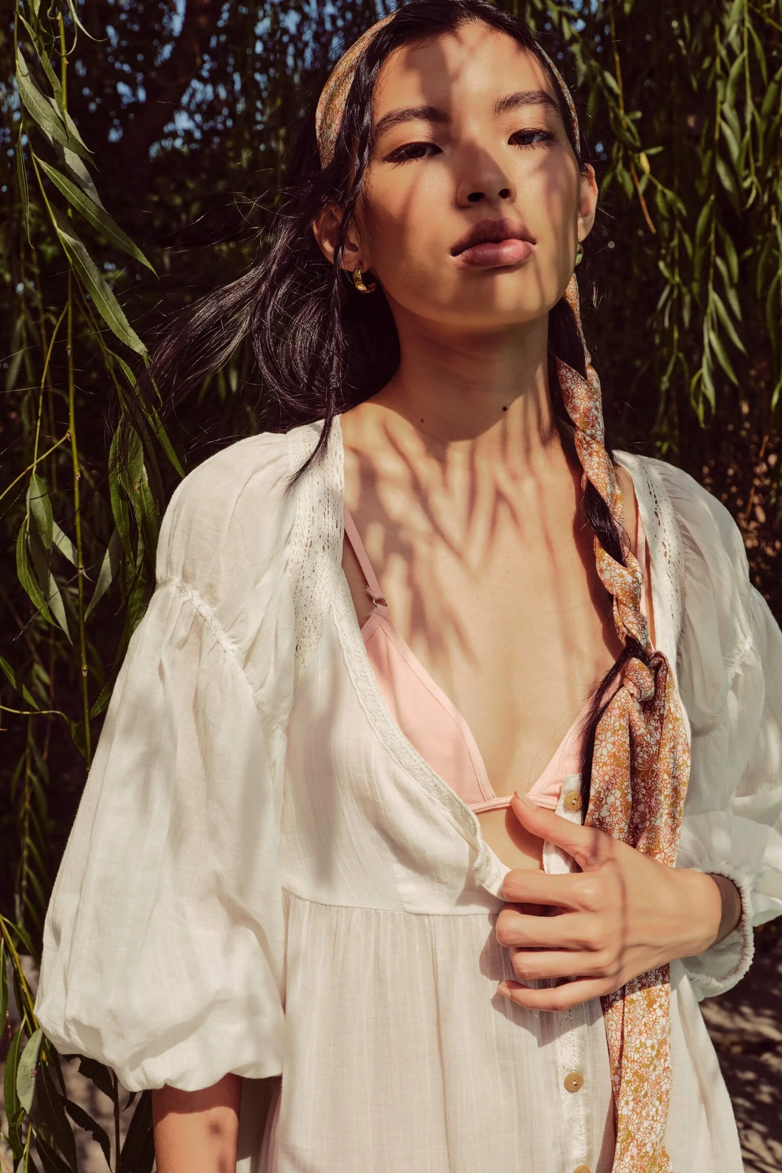 A woman standing outdoors in sunlight with shadows of leaves on her face and chest. She is wearing a light-colored, loose-fitting dress and a pink bra underneath. She has a patterned scarf tied around her hair and is holding the front of her dress wi