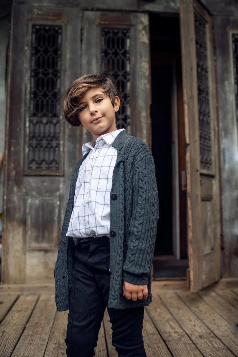 A boy standing on a wooden porch in front of an old, weathered wooden building with decorative windows, wearing a white checked shirt, a dark cardigan, and black pants.