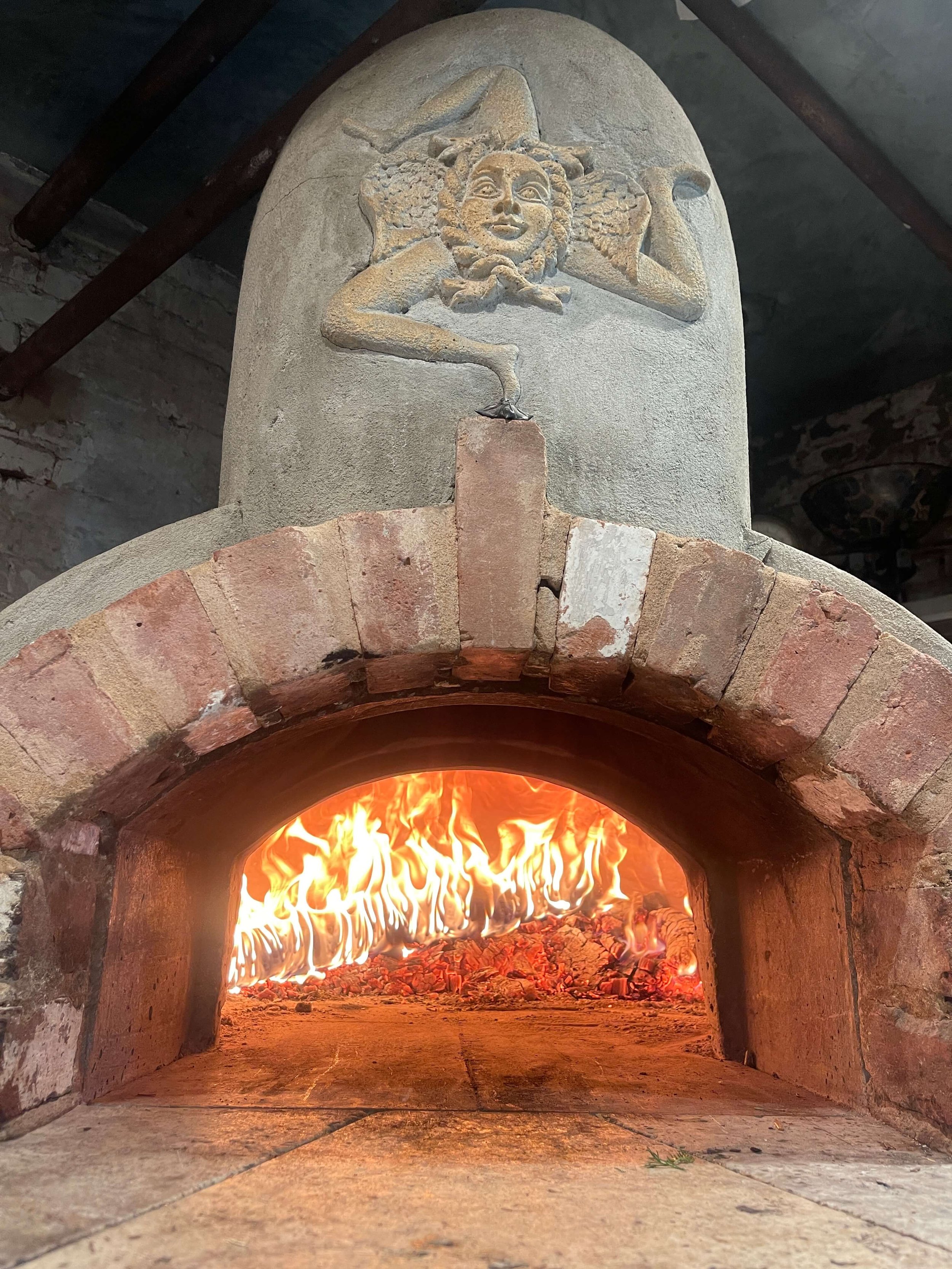 A traditional brick wood-fired oven with a fire burning inside. Above the oven, there is a sculpture of a smiling woman with curly hair and a lively expression, made of stone.