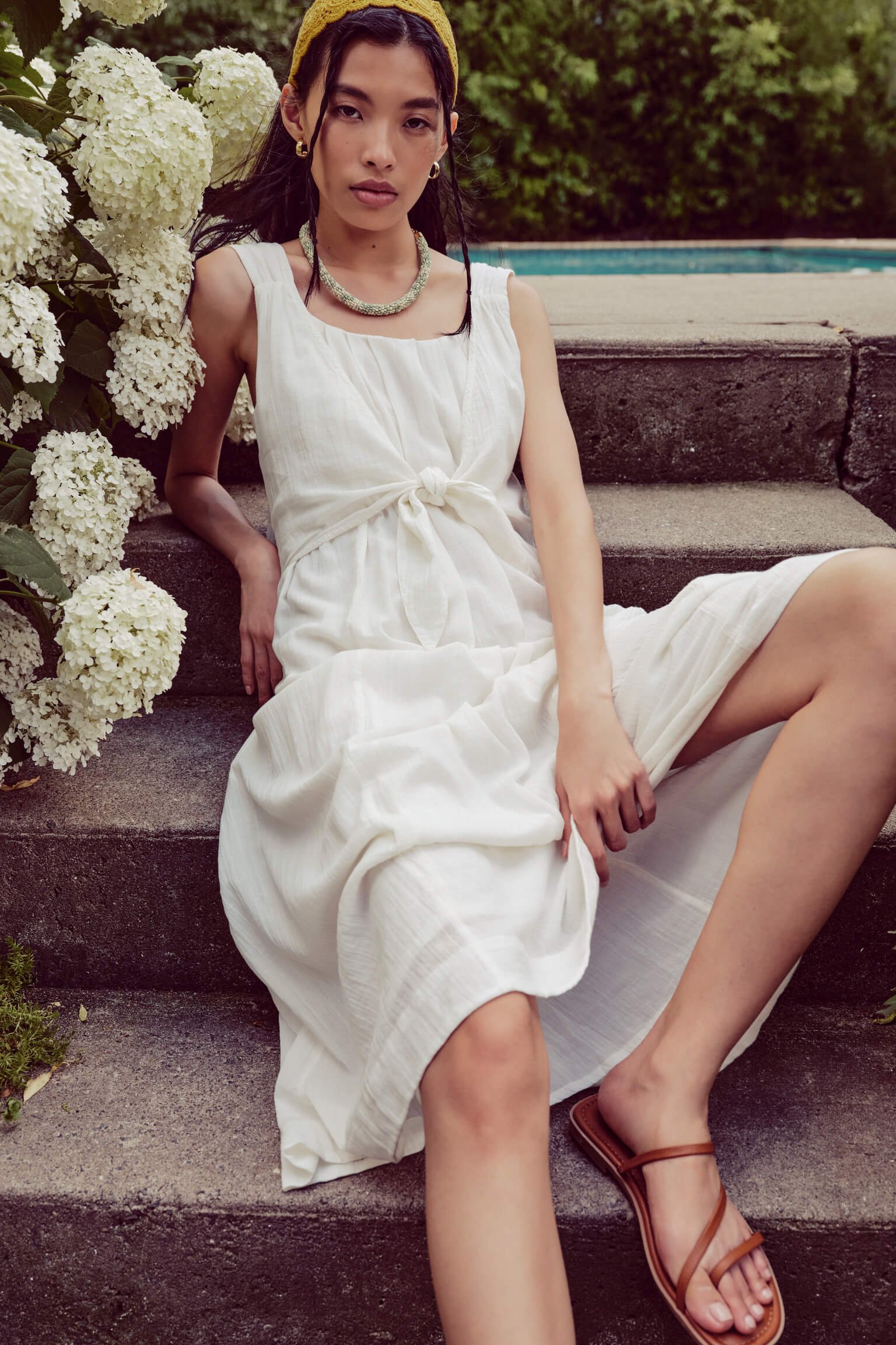 A woman with dark hair and gold jewelry, wearing a white dress and a yellow headband, sitting on outdoor stone steps with white hydrangea flowers and greenery in the background.