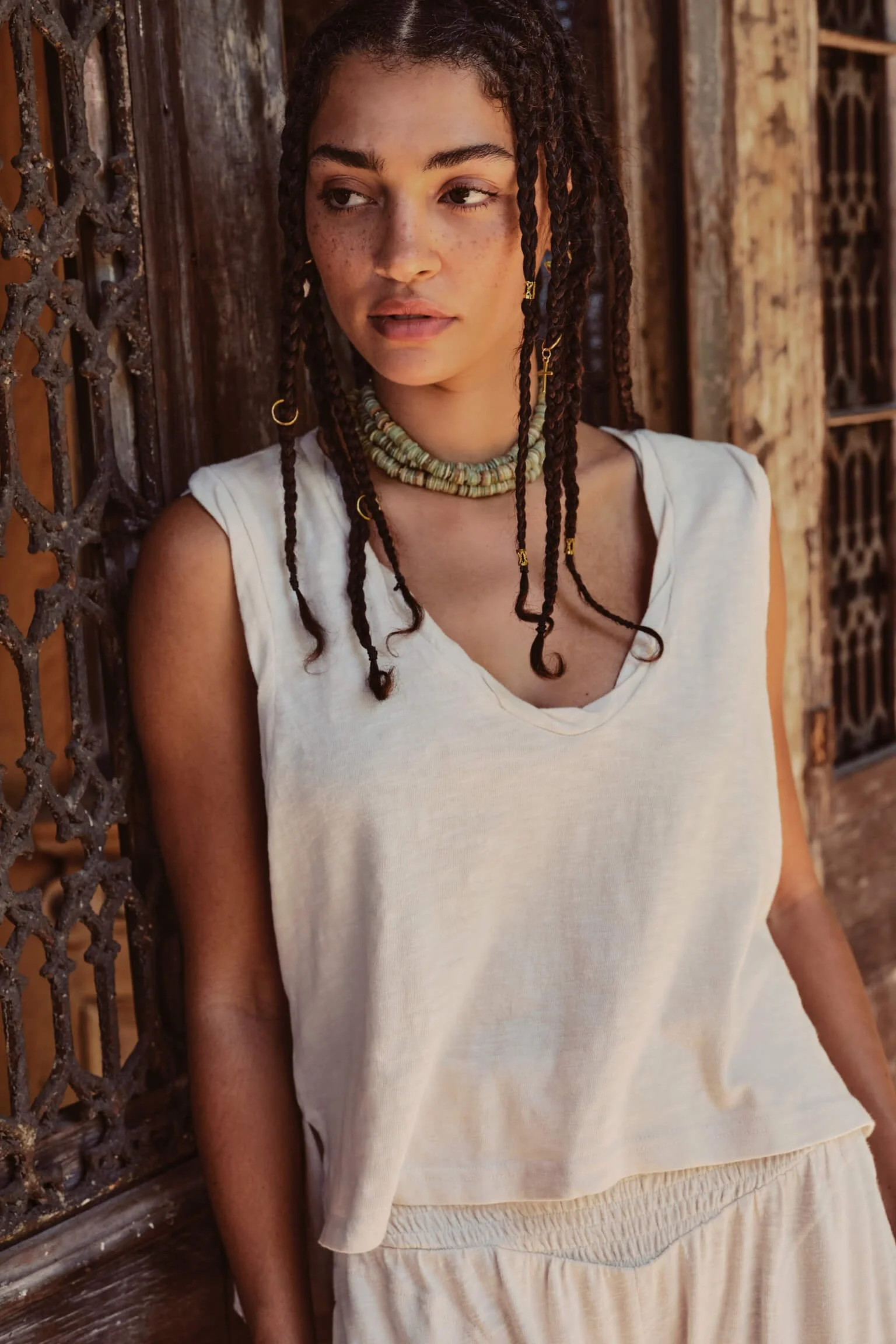 A young woman with braided hair wearing a white sleeveless top and beige pants, standing against a rustic wooden and iron fence.