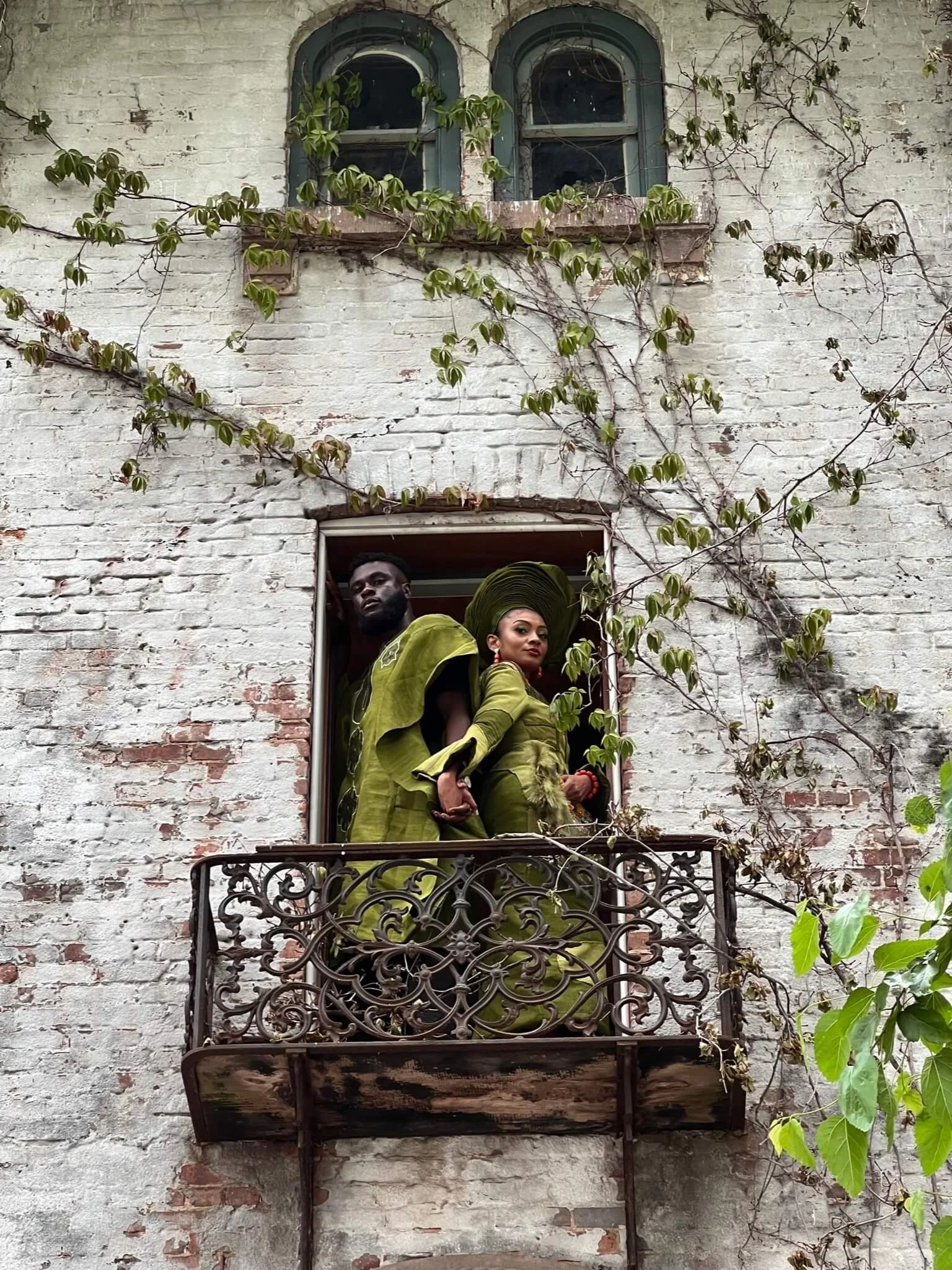 Two individuals dressed in green traditional attire standing on a small iron balcony outside an old white brick building with vines growing on the wall.