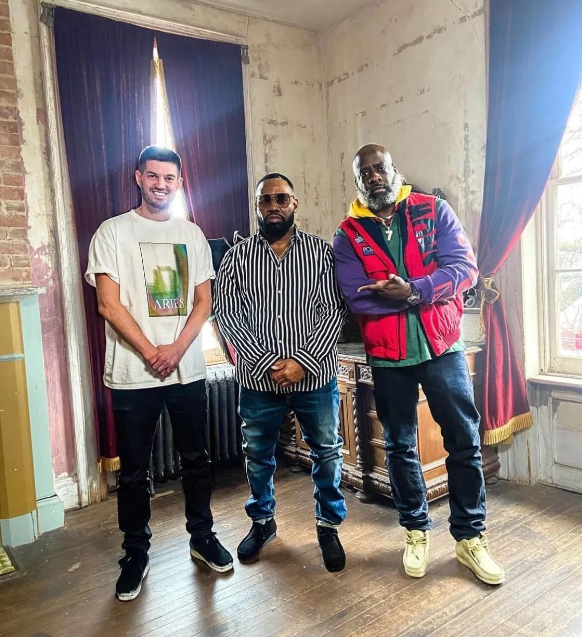 Three men standing indoors in front of a window with curtains. The man on the left is wearing a white T-shirt with a colorful design, the man in the middle is wearing a black and white striped shirt and sunglasses, and the man on the right is wearing