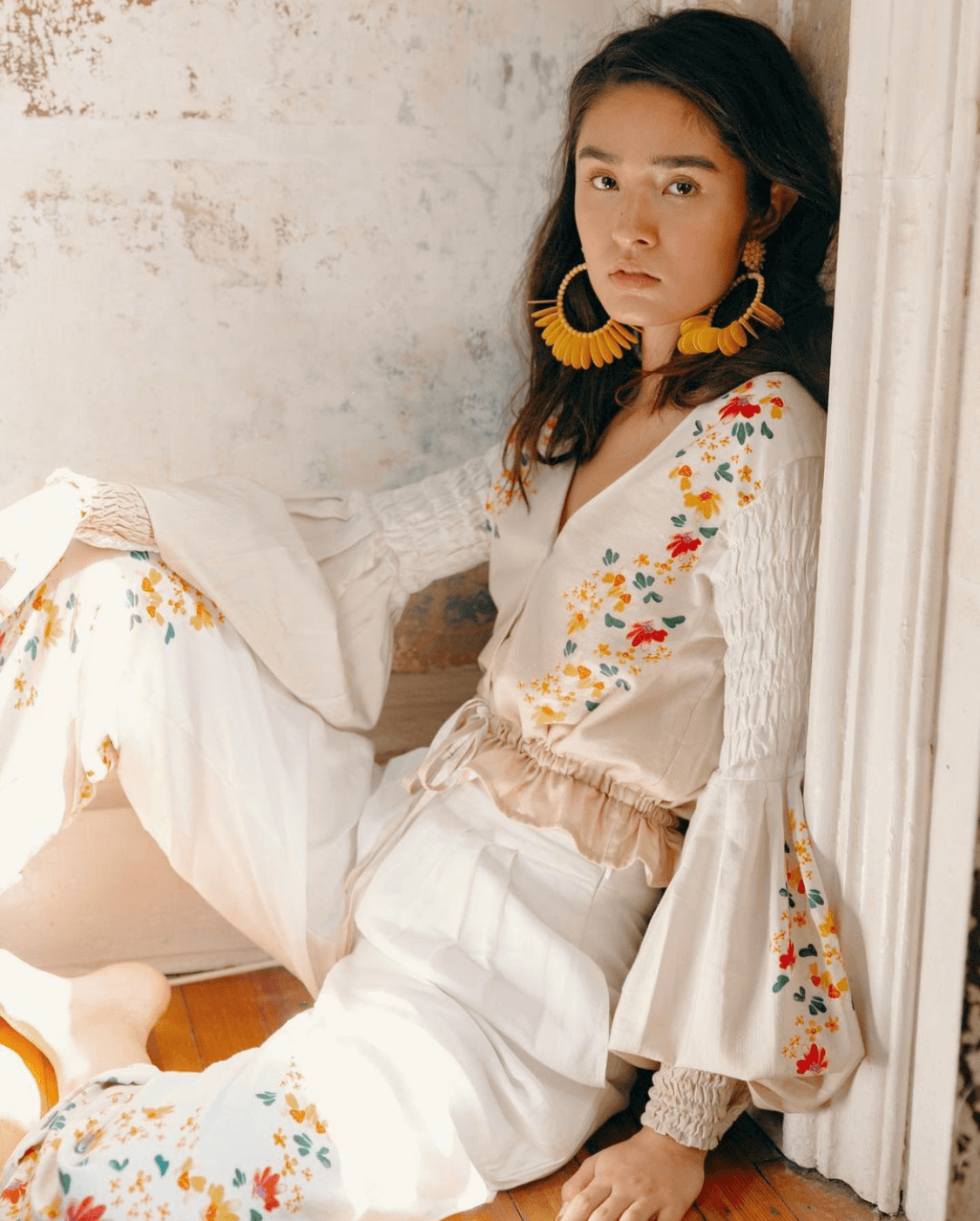 A woman with dark hair, wearing large yellow and black hoop earrings, and a cream-colored embroidered dress with colorful floral patterns, sitting on the floor and leaning against a wall.