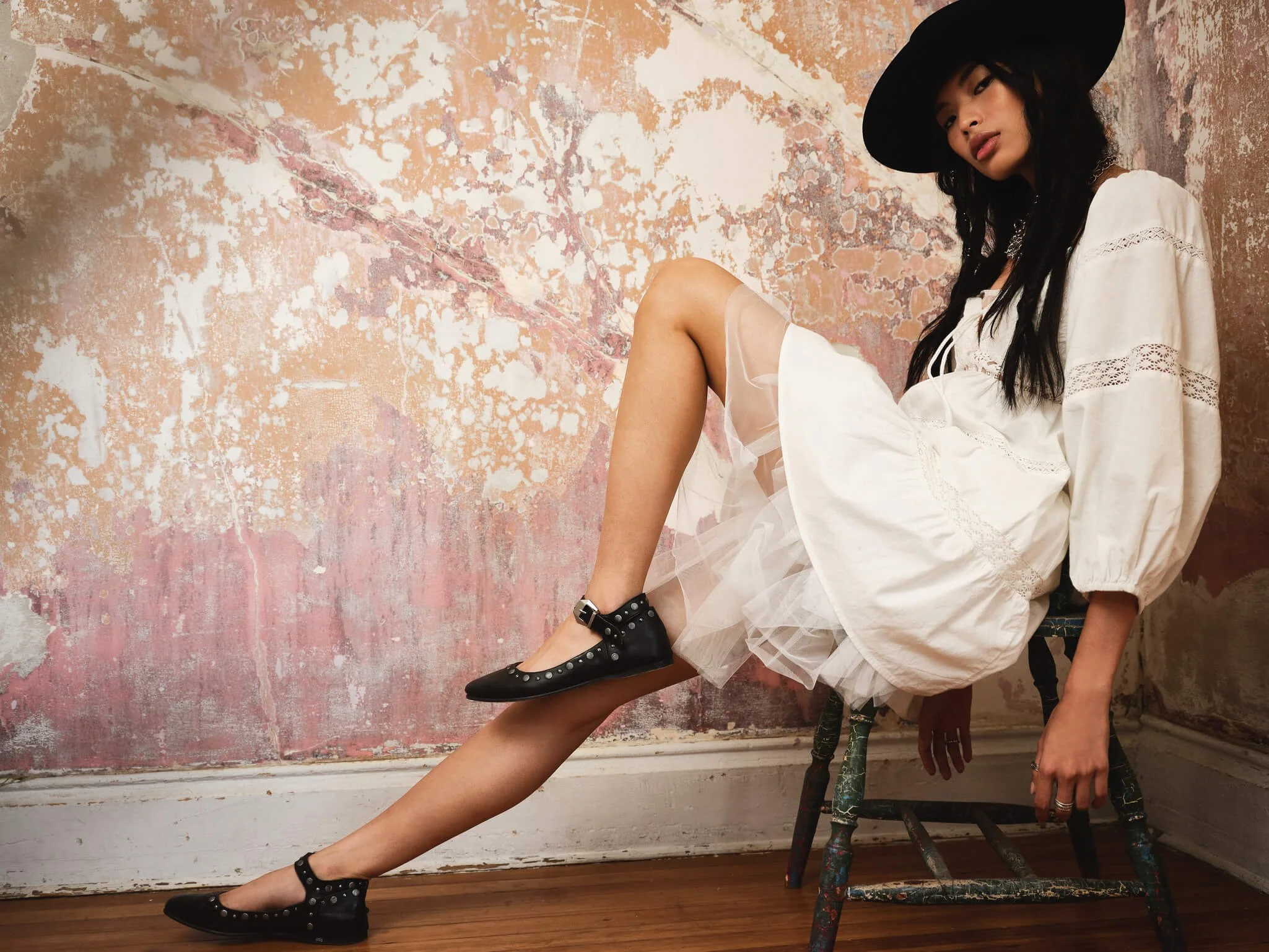 A woman wearing a white dress with lace details, a large black hat, and black flats with silver studs, sitting on a vintage chair against a distressed, multicolored wall.