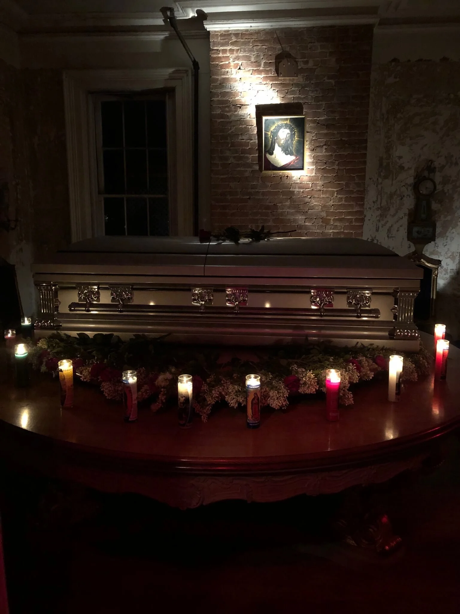 A casket sits on a wooden table surrounded by lit candles and floral arrangements, with a brick wall and religious artwork in the background.
