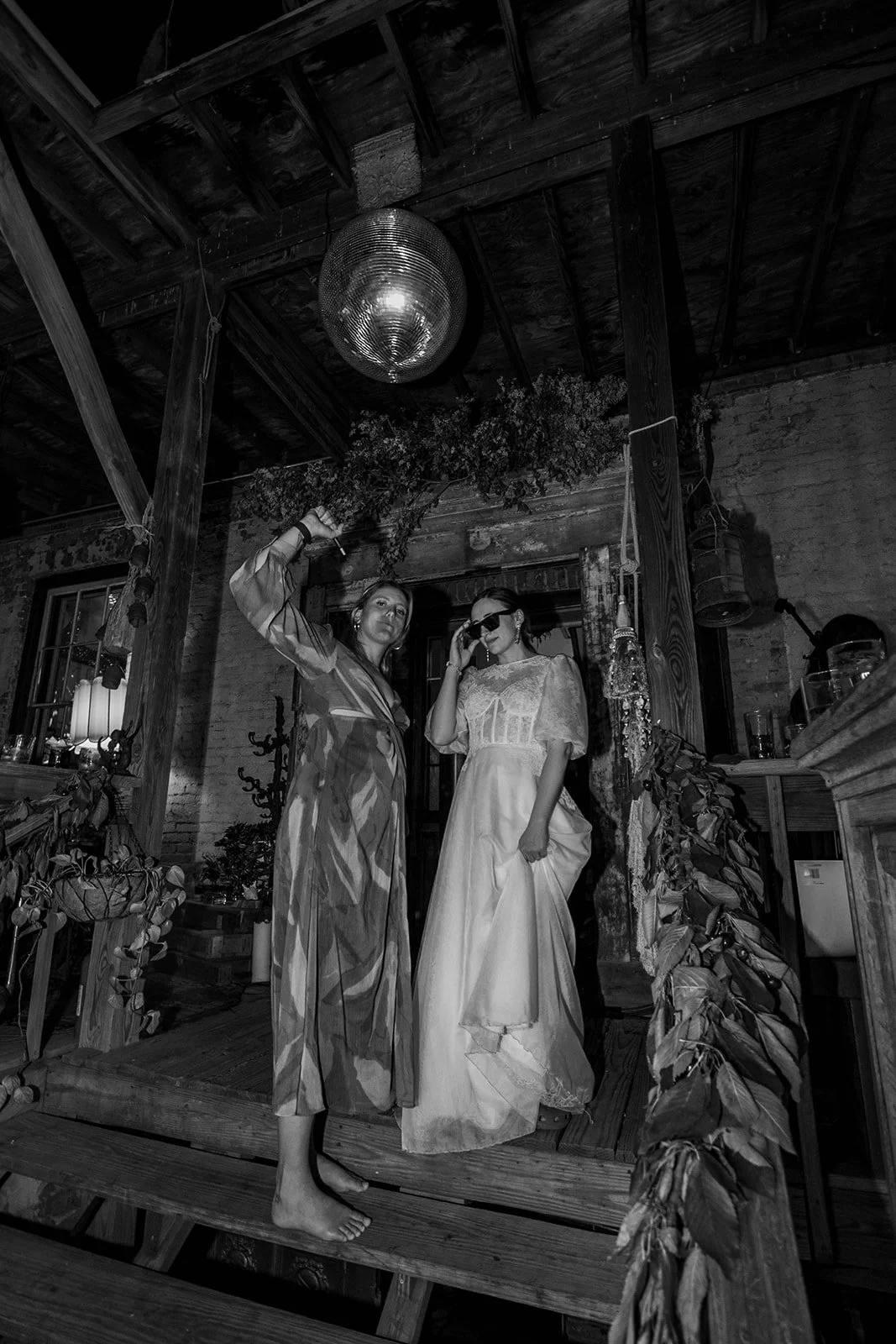 Two women, one in a patterned dress and the other in a wedding gown, posing backstage at a rustic event venue with wood beams, plants, and a disco ball overhead.