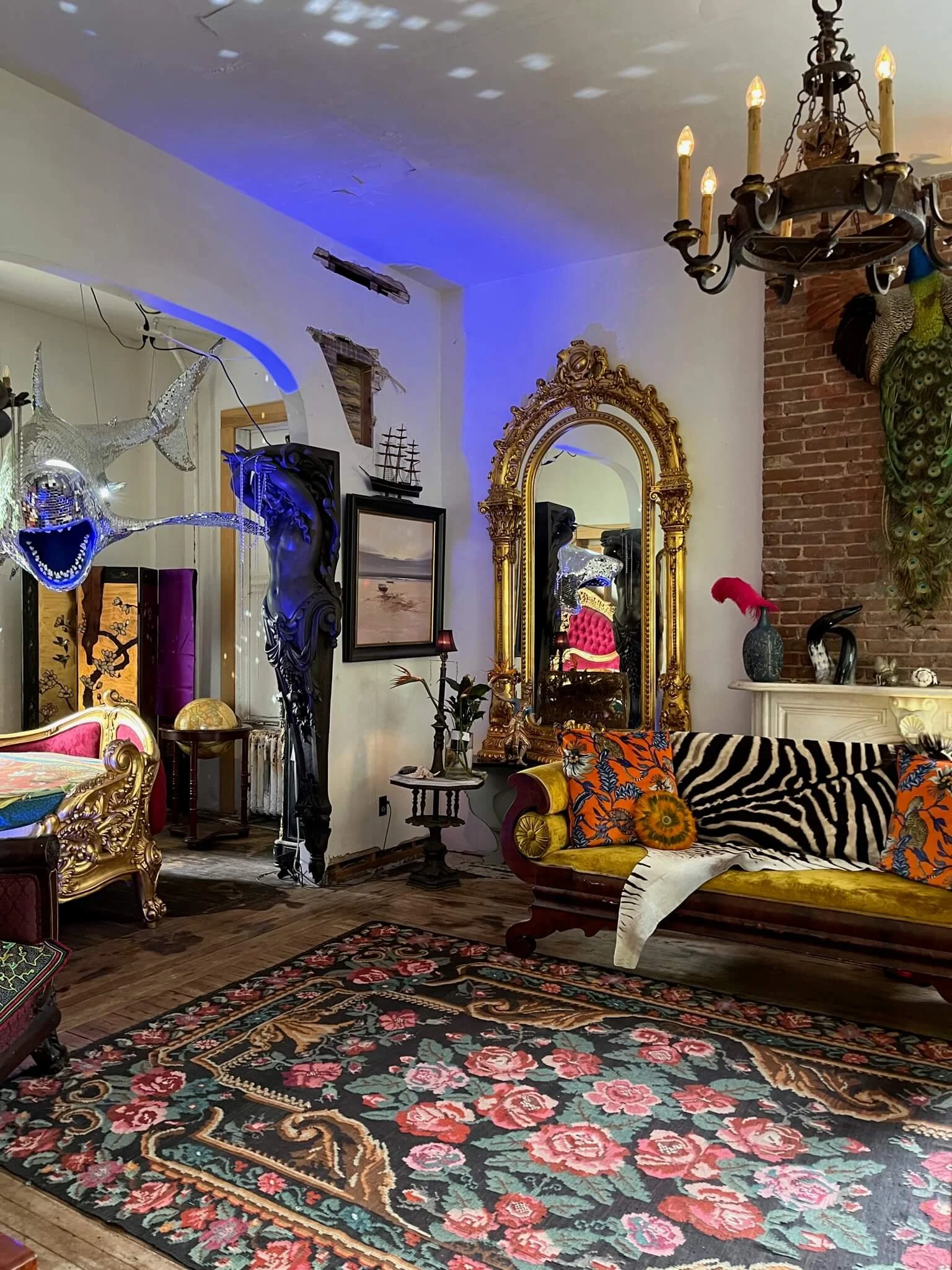 An eclectic living room with a floral area rug, ornate gold-framed mirror, vintage sofa with colorful pillows, a large peacock and peacock feathers art piece, and various decorative objects including a ship model, vases, and art on the walls.