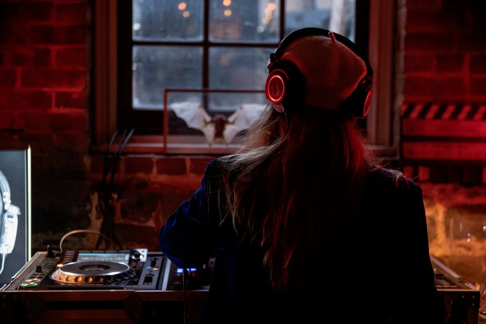 A female DJ wearing headphones and a beanie, seen from behind, performing in a dimly lit venue with red and blue lighting, brick walls, and a window with a moth inside.