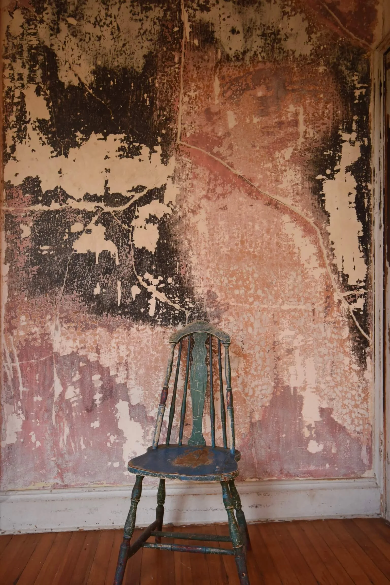 An old, worn wooden chair with peeling paint is placed in front of a distressed, textured wall with peeling paint and patches of different colors in a rustic room with wooden floorboards.