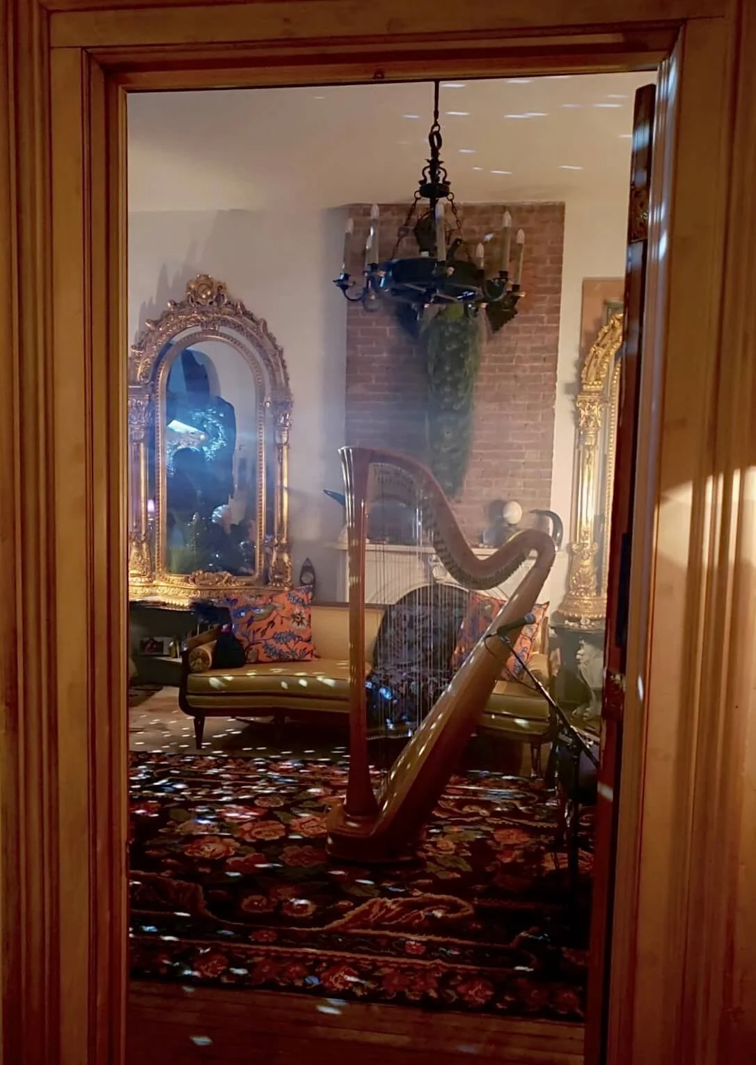 View of an elegant room seen through a glass door, featuring a harp, ornate gold-framed mirrors, a chandelier, a vintage sofa with colorful pillows, and a patterned rug.