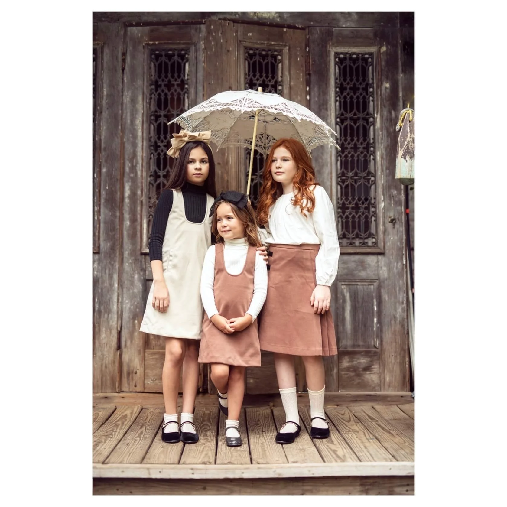 Three young girls standing on a wooden porch in front of a rustic wooden door, with one holding a lace parasol. They are dressed in vintage-style clothing with neutral and earthy tones.