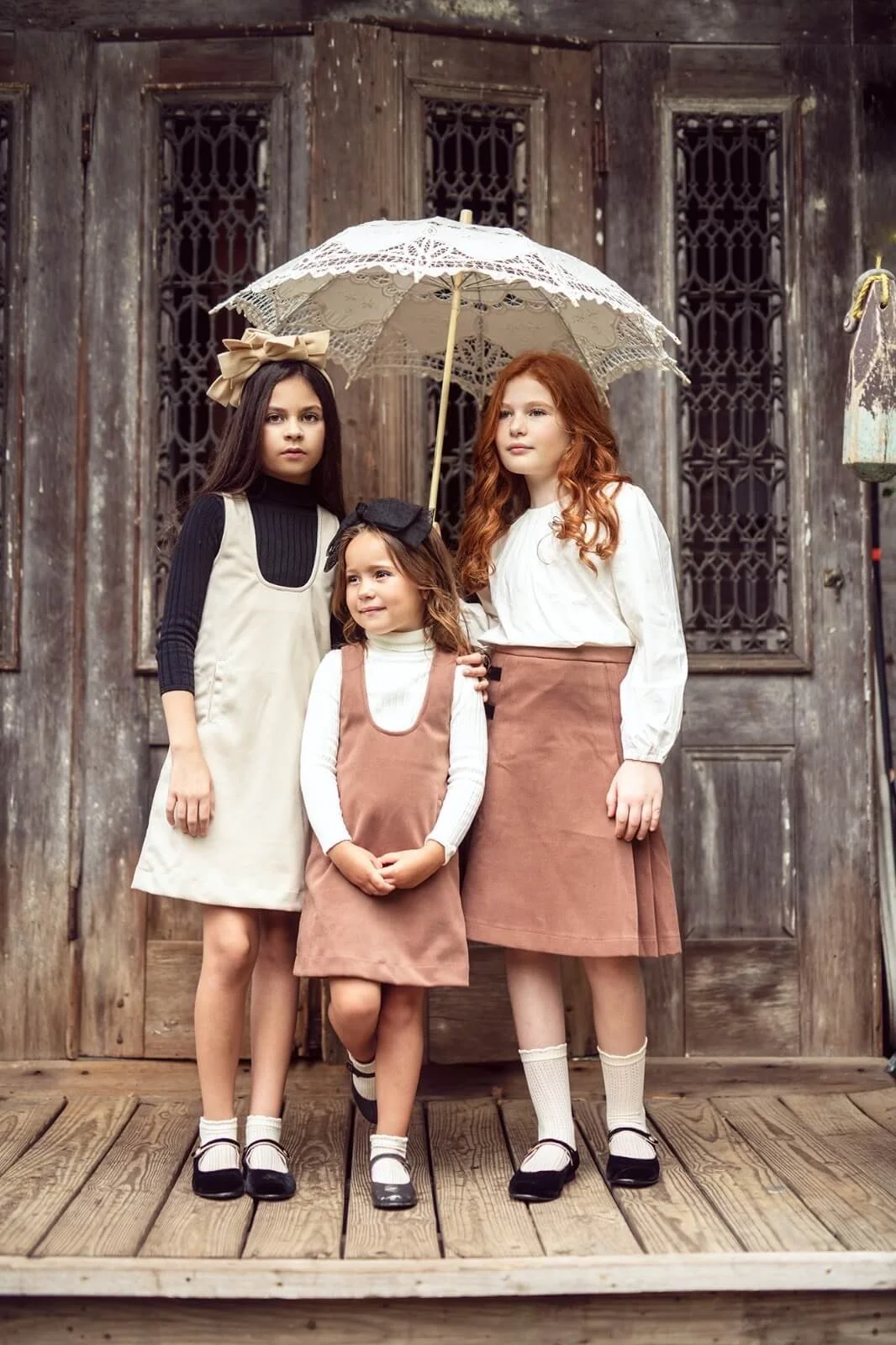 Three young girls standing on a wooden porch in vintage-style clothing, holding a lace umbrella, in front of an old wooden door with iron bars.