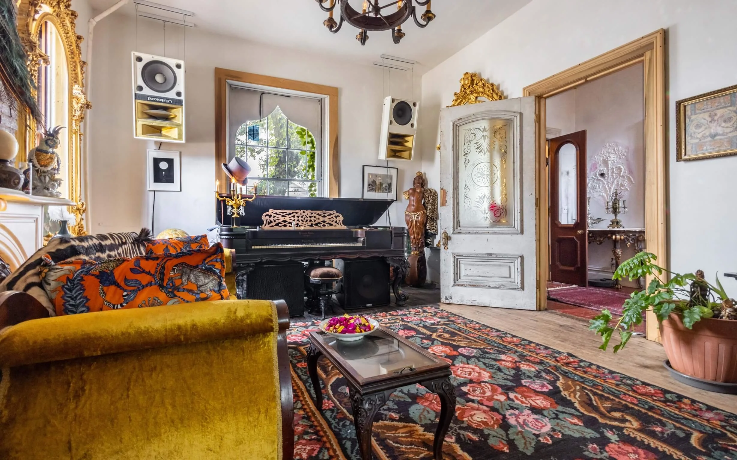 Living room with vintage decor, including a black grand piano, colorful patterned rugs, plush yellow velvet sofa, ornate gold-framed mirror, and tropical potted plants.