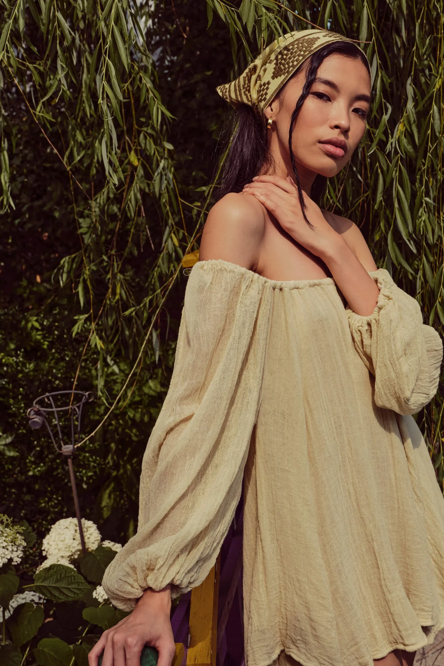 A woman in a yellow off-shoulder dress, wearing a patterned headscarf, standing outdoors near green foliage and white flowers.