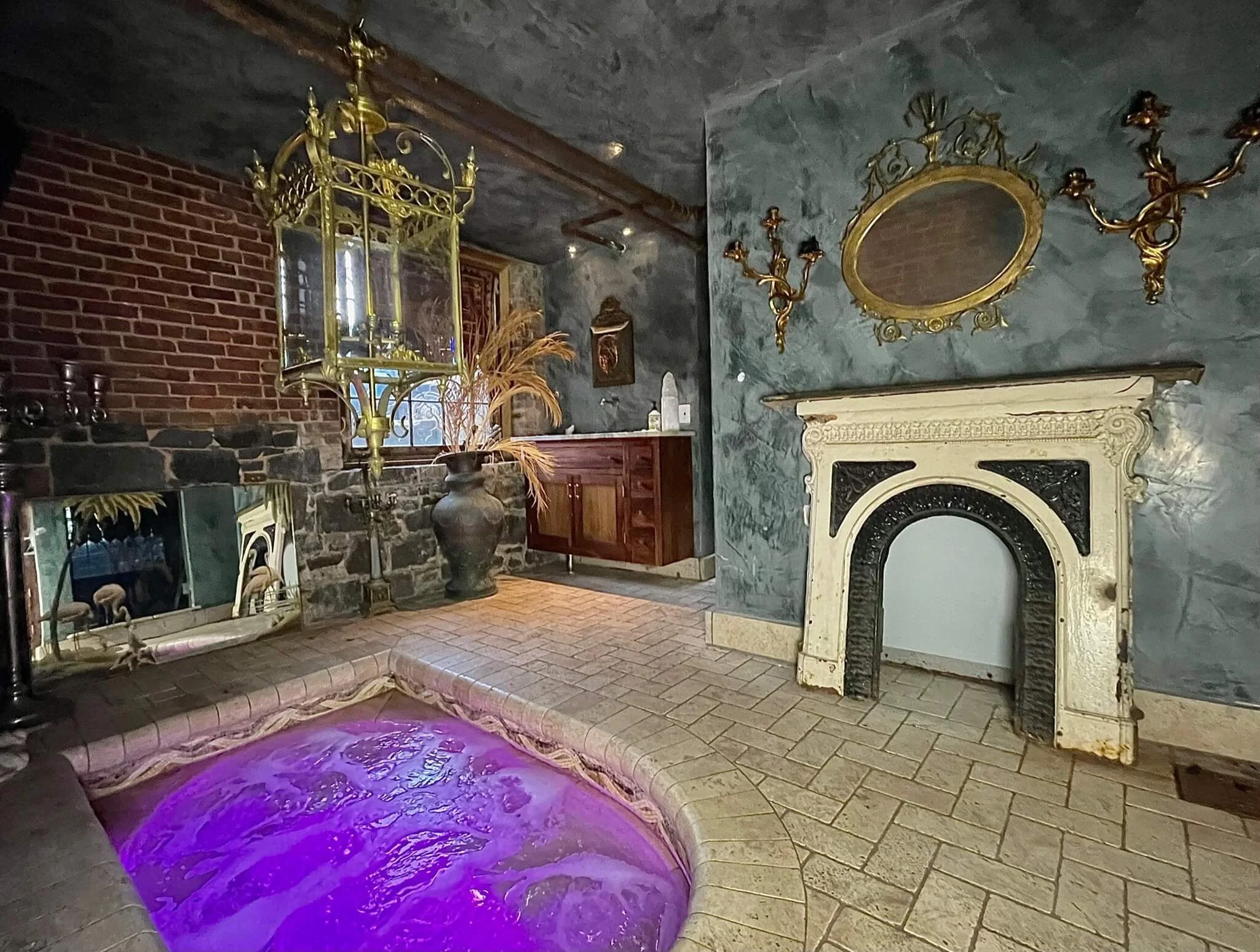 Indoor spa room with a lit purple hot tub, stone and brick walls, a vintage fireplace, ornate gold wall sconces, a large mirror, and decorative antique furnishings.