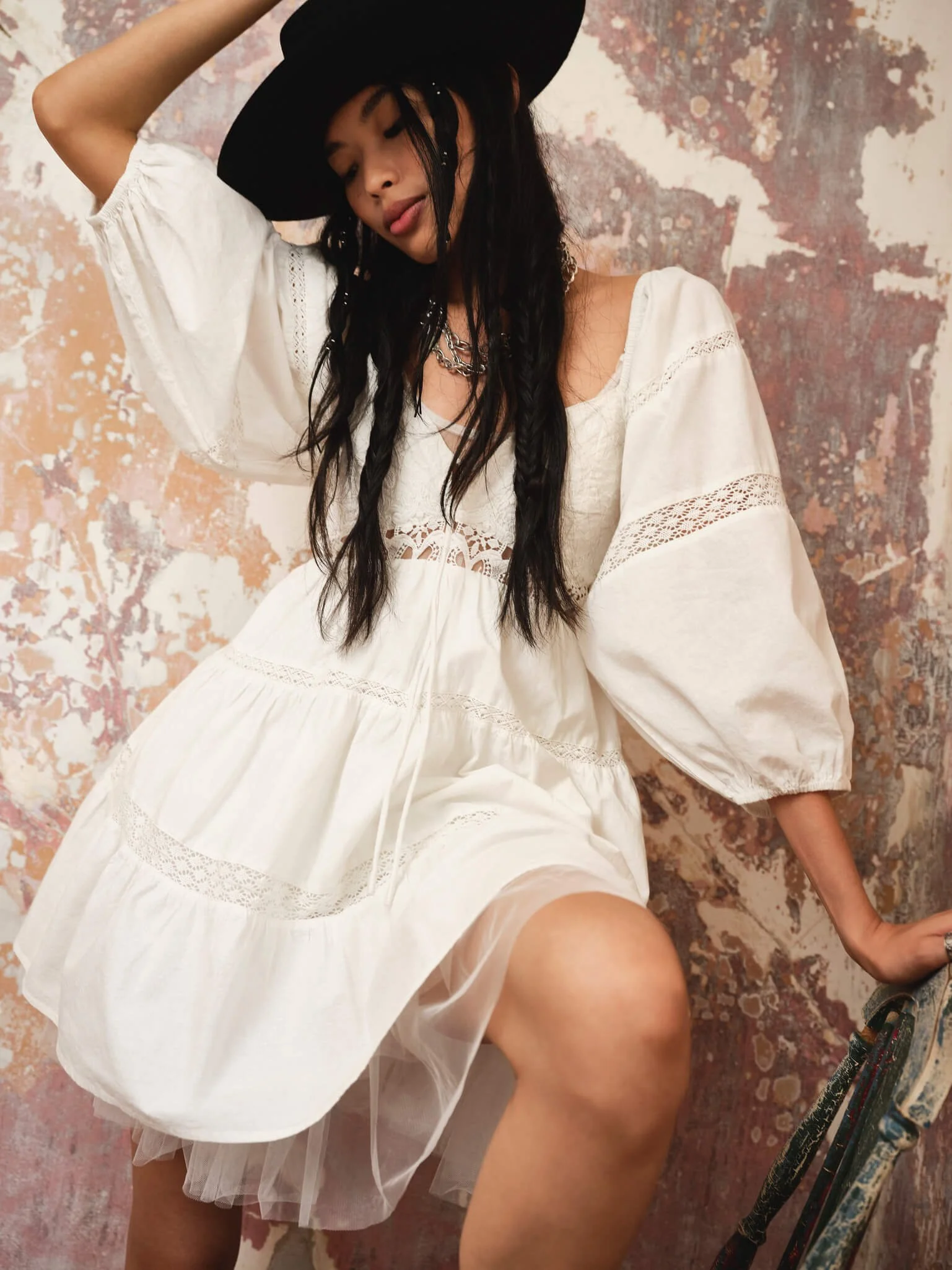 A woman with long dark hair wearing a wide-brimmed black hat and a cream dress with lace trim, sitting against a textured, painted wall.