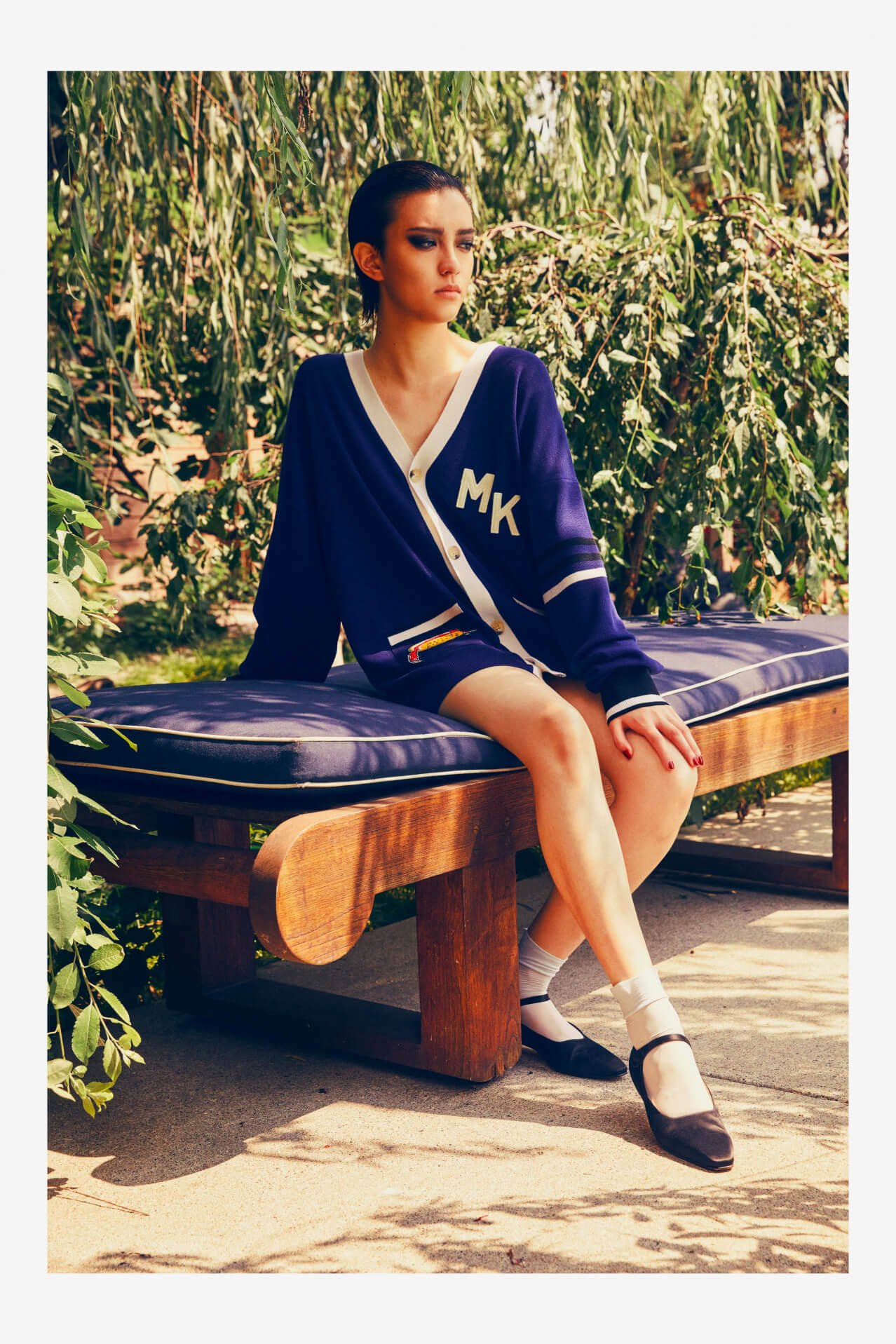 A young woman with dark hair and makeup sitting on a wooden bench outdoors, wearing a navy blue cardigan with white accents and initials 'MK', beige socks, black shoes, and shorts, surrounded by greenery and sunlight.