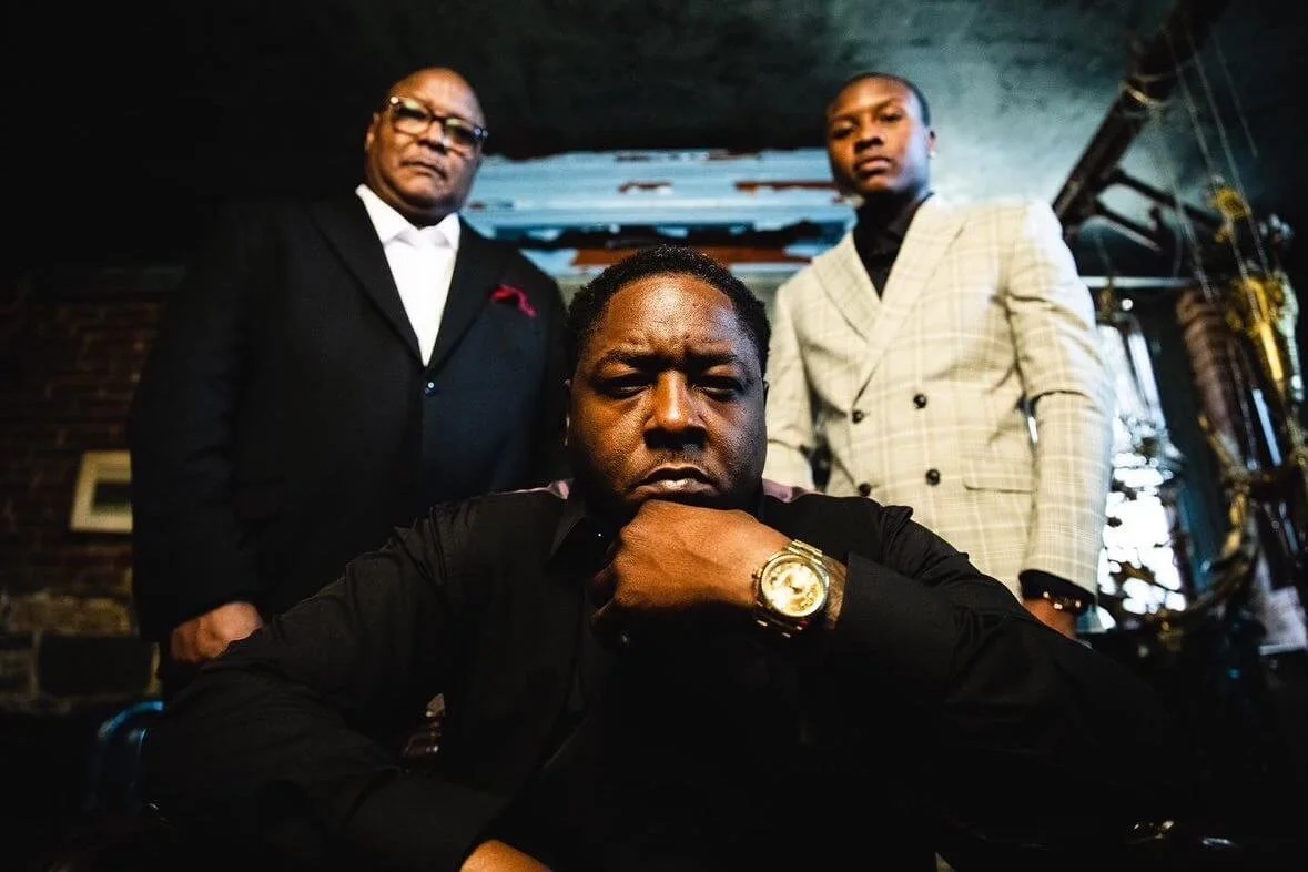 Three men in stylish suits posing in a dimly lit room with exposed brick walls and decorative lighting, with one man sitting in front and two standing behind him.