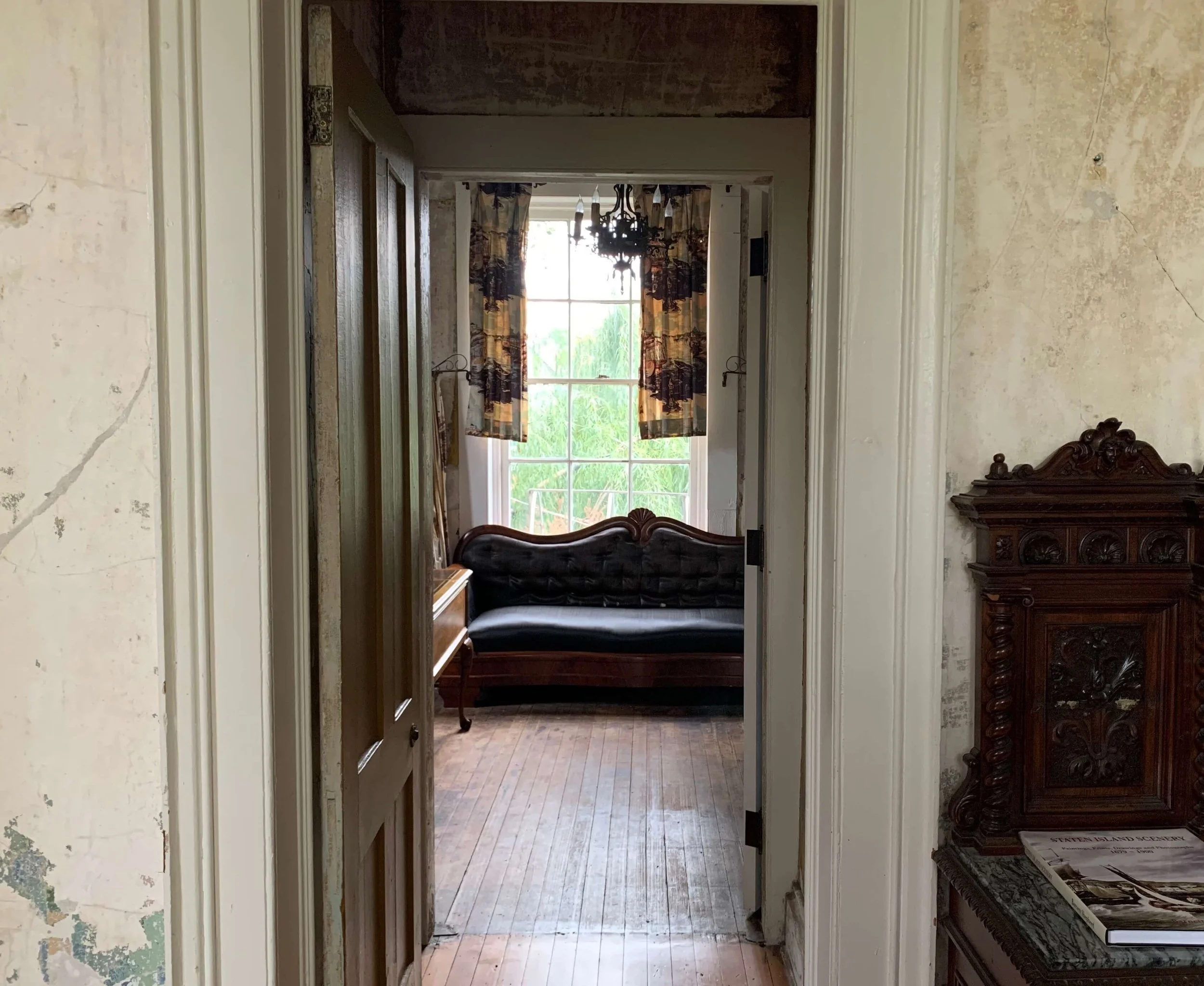  View of a vintage interior room through an open doorway, featuring a black velvet antique sofa, a large window with patterned curtains, and wooden furniture with intricate carvings.