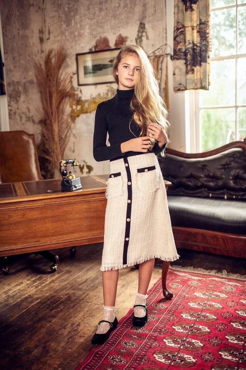 A young woman with long blond hair wearing a black turtleneck, cream-colored skirt with black accents, white socks, and black shoes standing in a vintage-style living room by a wooden desk and an antique rotary phone.