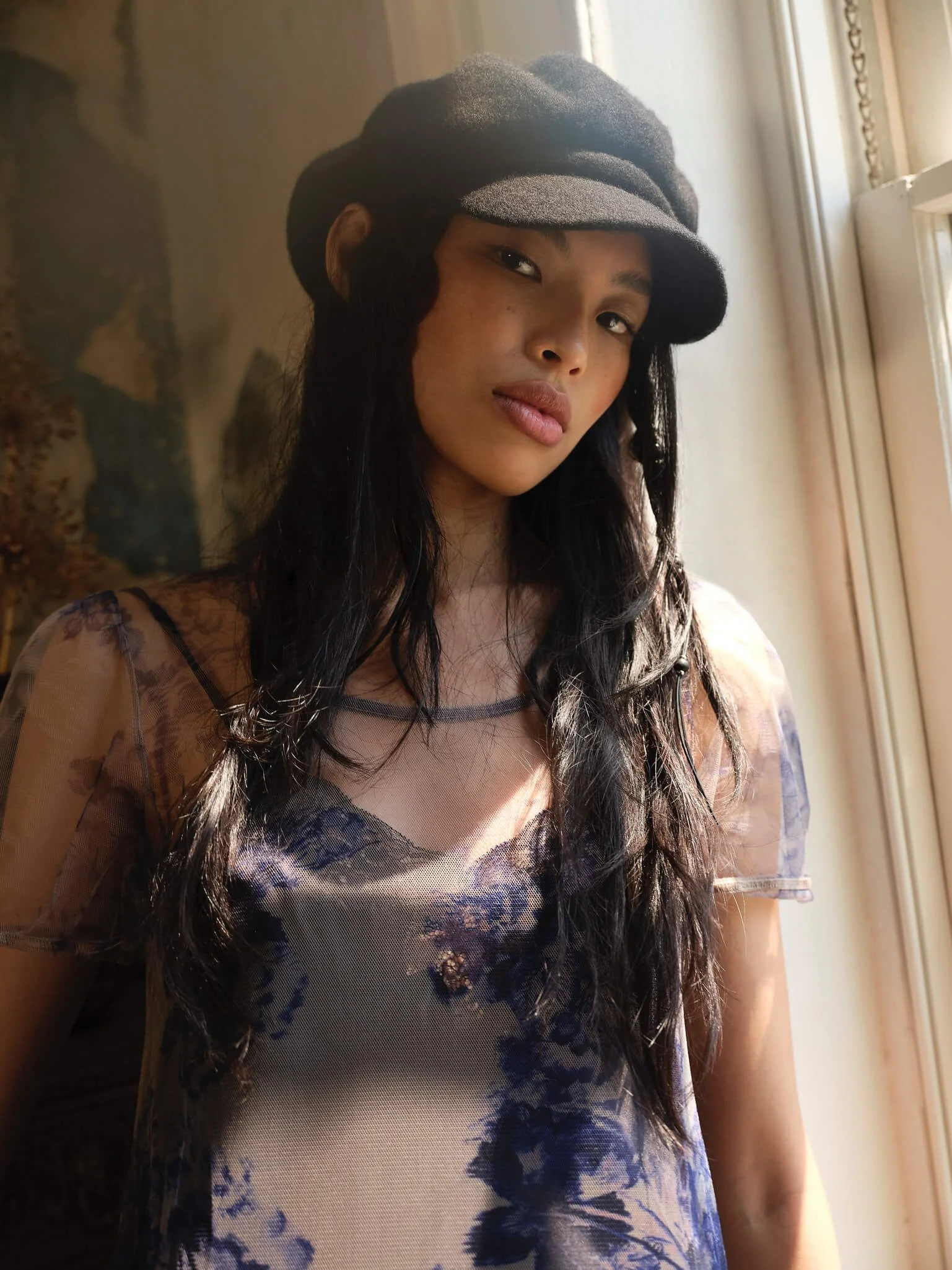 A woman with long dark hair wearing a black hat and a sheer floral top stands by a window, looking at the camera.