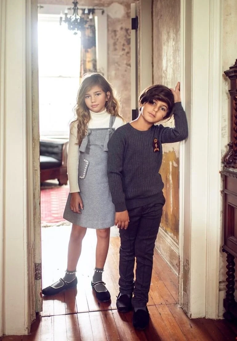 Two children, a girl and a boy, standing in a doorway indoors. The girl has long, wavy hair and is dressed in a white turtleneck and a gray dress. The boy has short hair and is wearing a dark sweater and pants, with one hand resting on the doorframe.