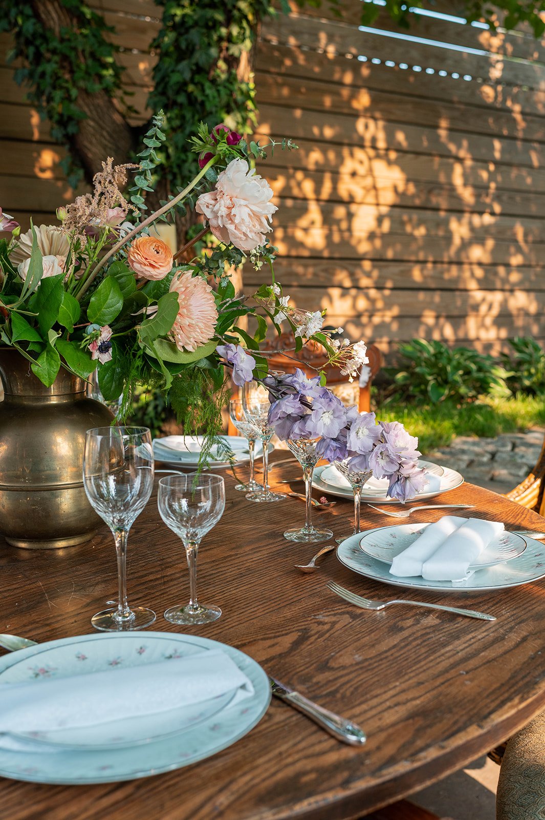 Elegant outdoor dining table decorated with a large floral centerpiece, wine glasses, and place settings with napkins, set against a wooden fence and greenery.