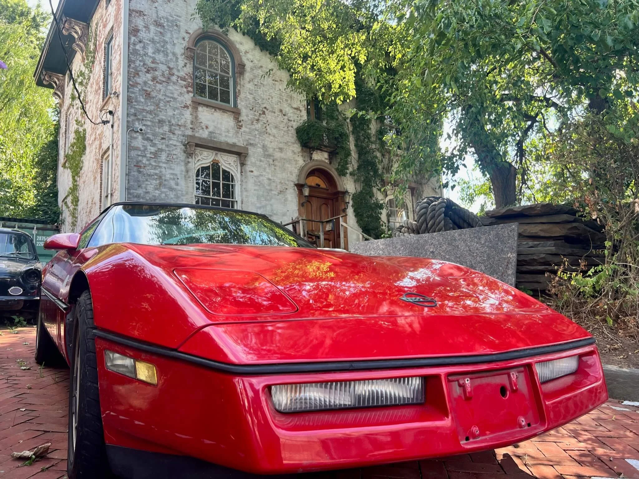 A red vintage sports car parked on a brick driveway in front of an old, weathered house surrounded by trees.
