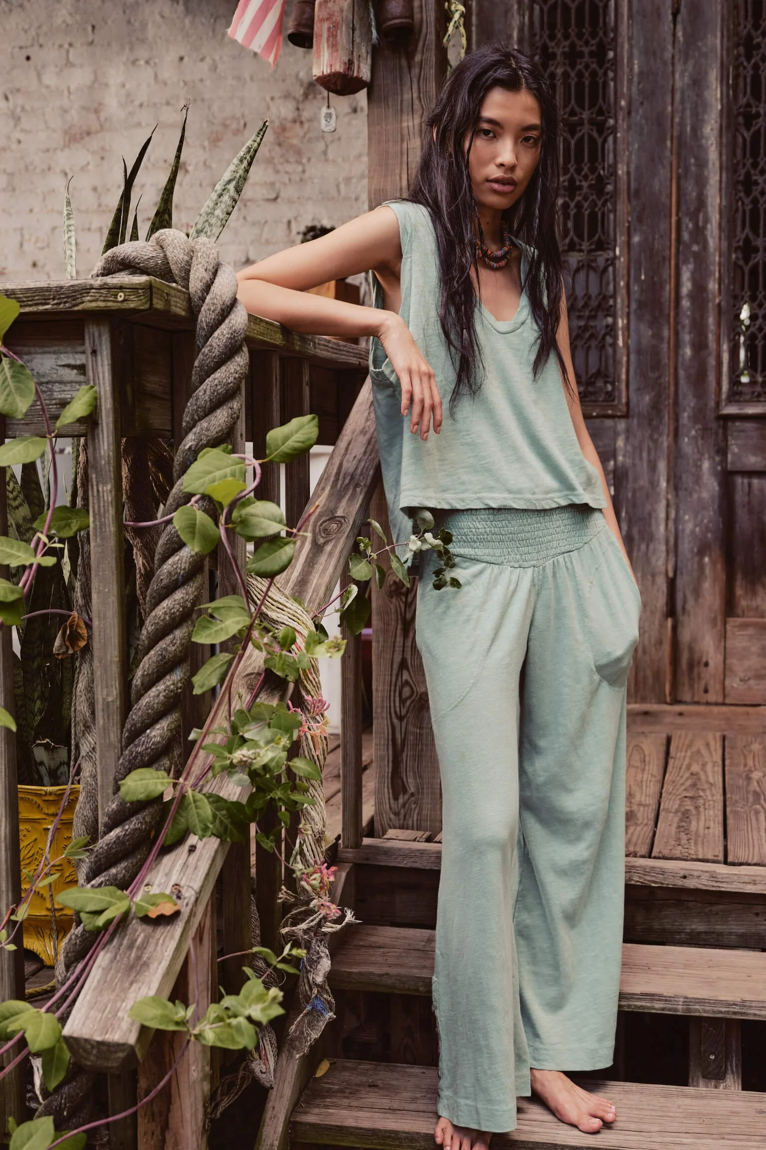 A woman with long dark hair wearing a light green sleeveless top and matching loose pants standing on a wooden staircase outdoors surrounded by plants and rustic wooden structures.