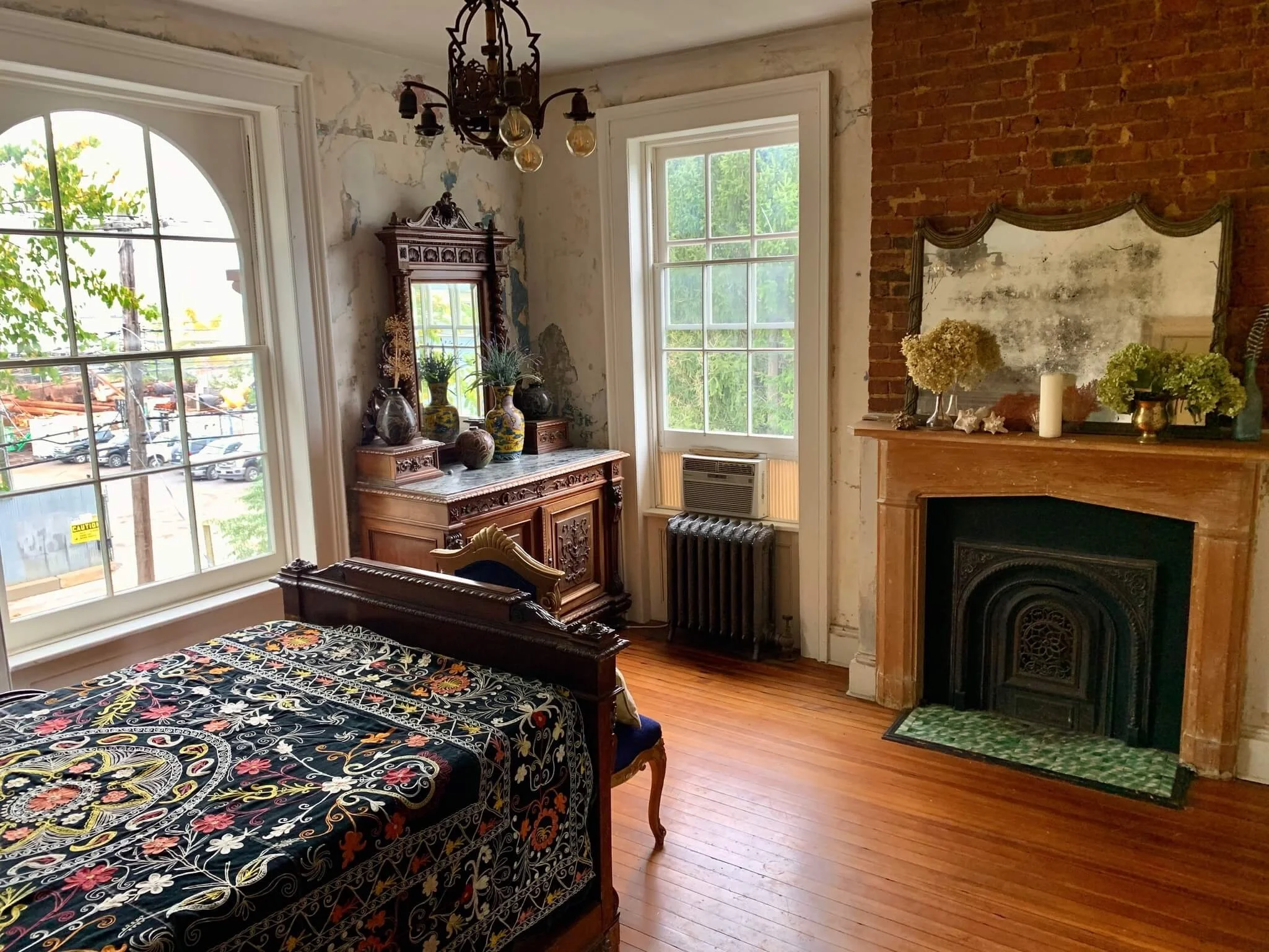A vintage bedroom with a wooden bed, floral bedspread, a large window, a fireplace with a mirror on top, and an ornate wooden dresser with vases and decorative items.