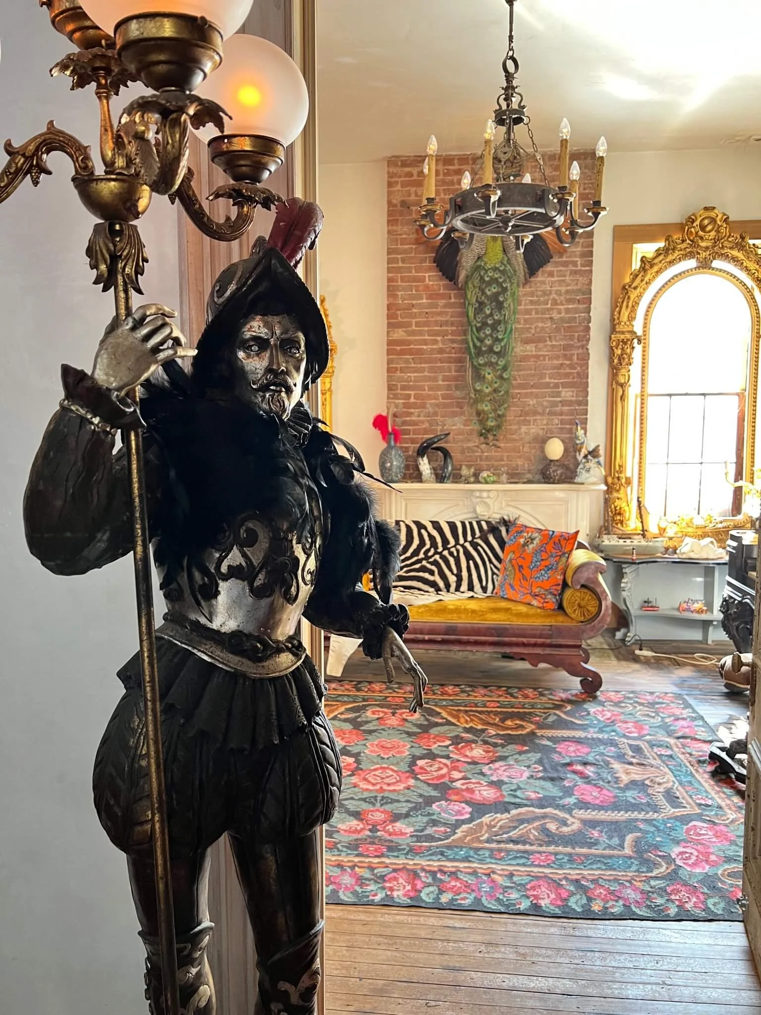 A detailed wooden or metal statue of a warrior holding a spear, positioned in a vintage, eclectic room with a ornate chandelier, colorful rug, patterned couch, and decorative wall art.