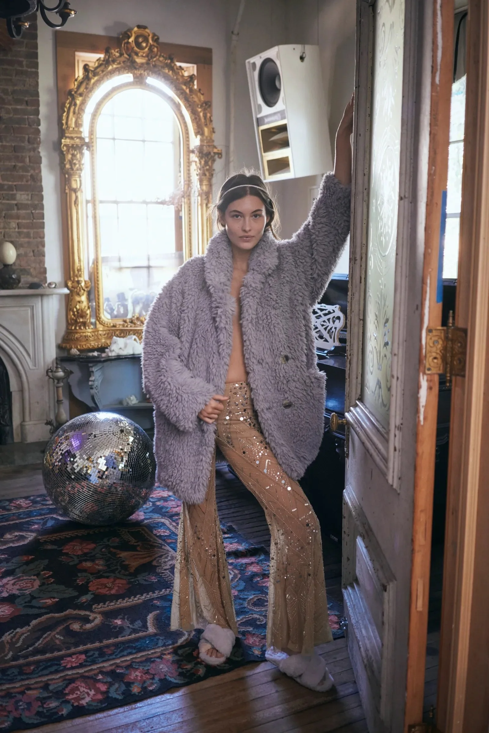A young woman posing inside a room with ornate decor, wearing a lavender faux fur coat and shiny beige pants with sequins, standing near a partially opened door.