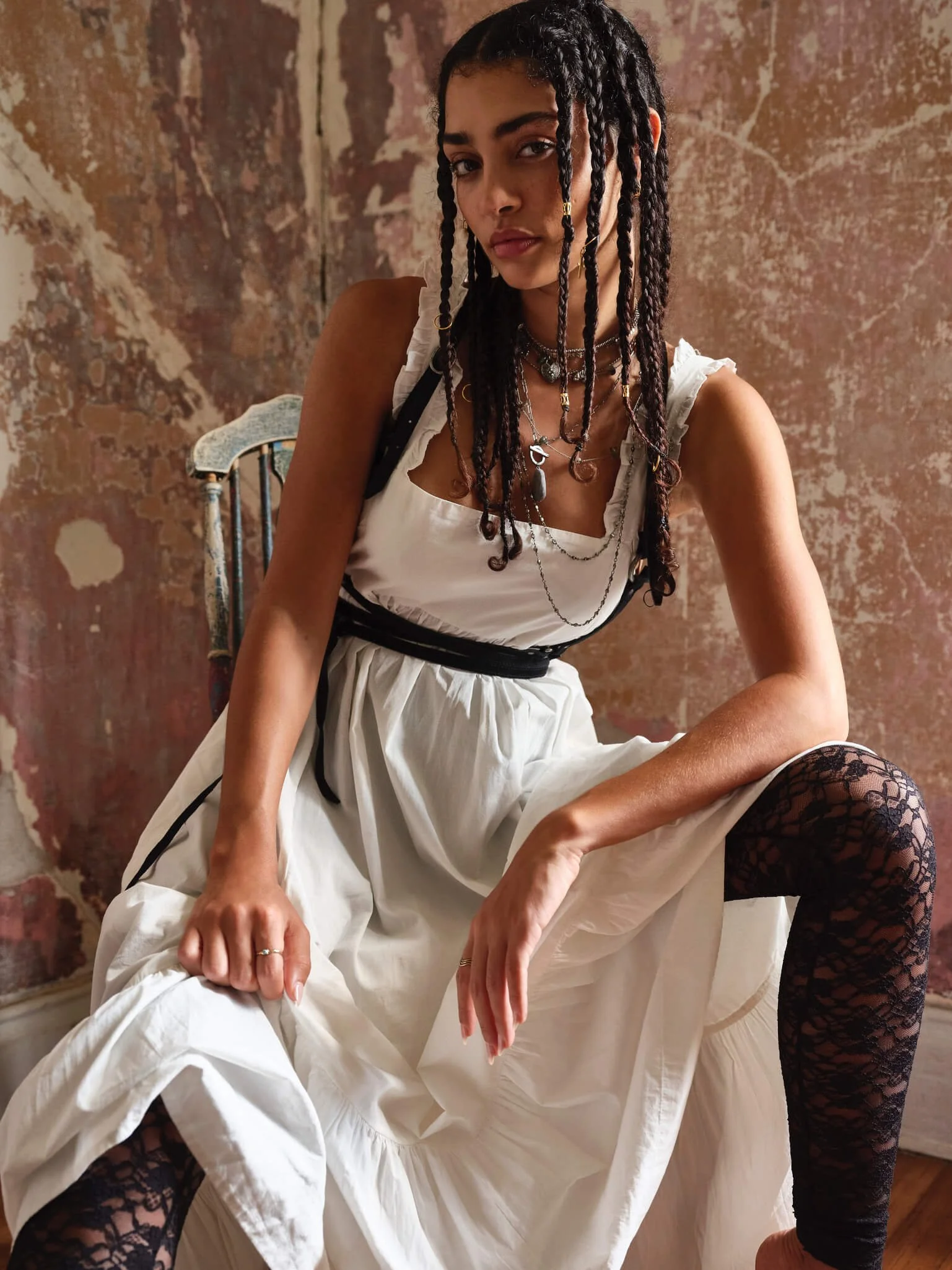 A young woman with long braided hair and multiple necklaces, wearing a white dress with black lace stockings, sits on a wooden chair in front of a distressed wall.
