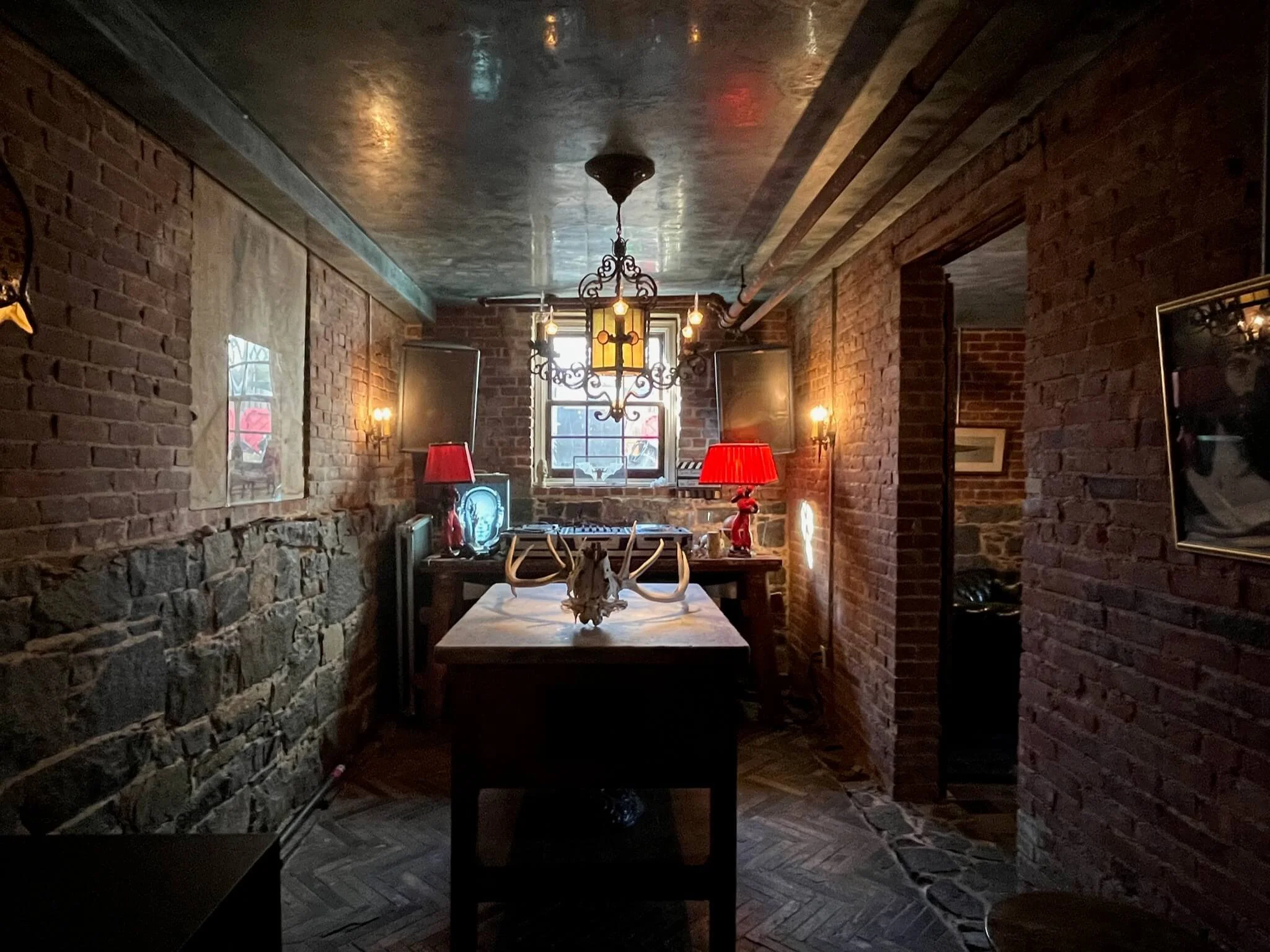 Dimly lit room with brick walls, stone accents, and a wooden table in the center. The table has a deer skull with antlers. Red lamps and a chandelier hang from the ceiling, and a window in the background lets in some natural light. There is equipment