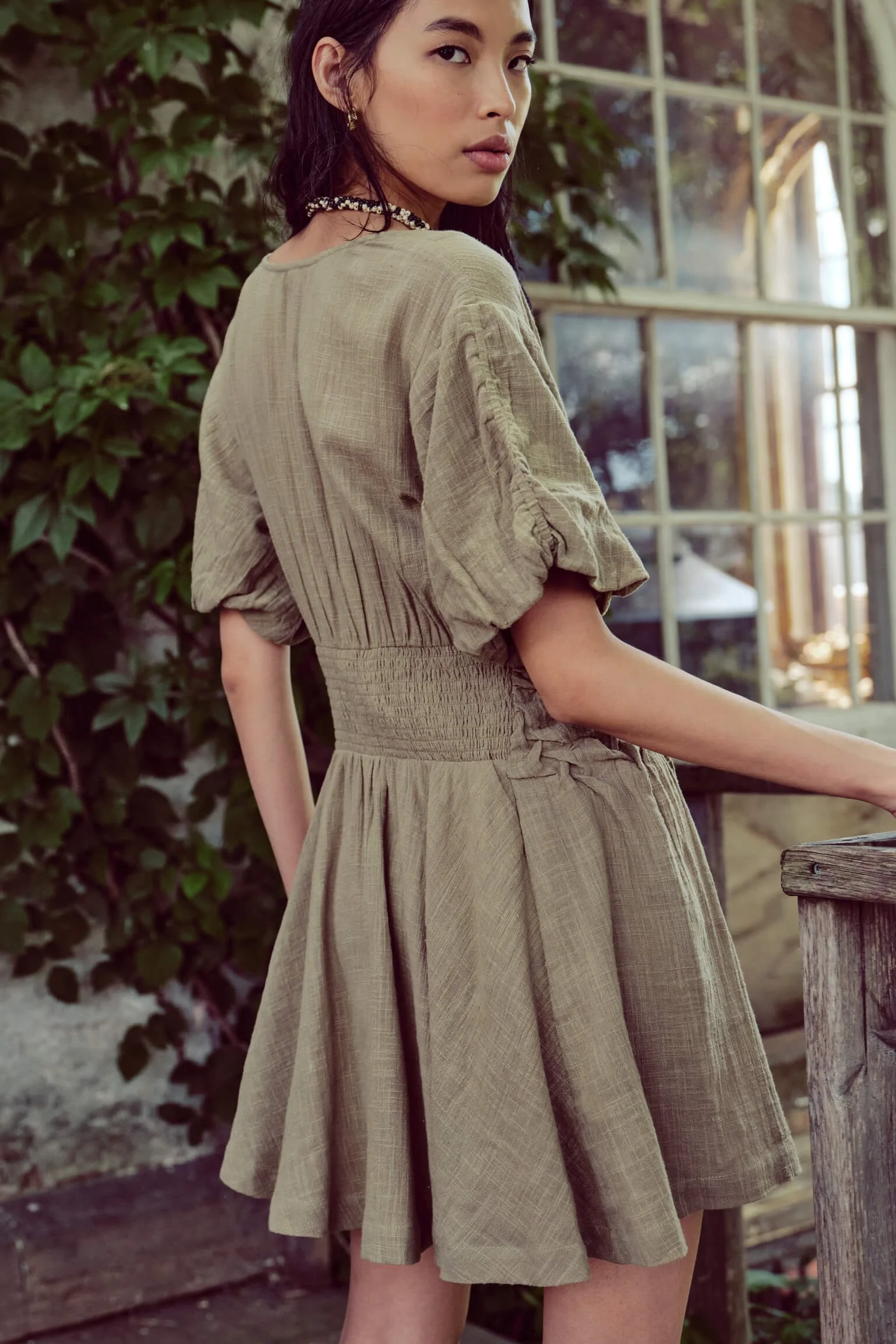 A woman with dark hair wears a beige dress with puffed sleeves and a cinched waist, standing indoors near a window with greenery outside.