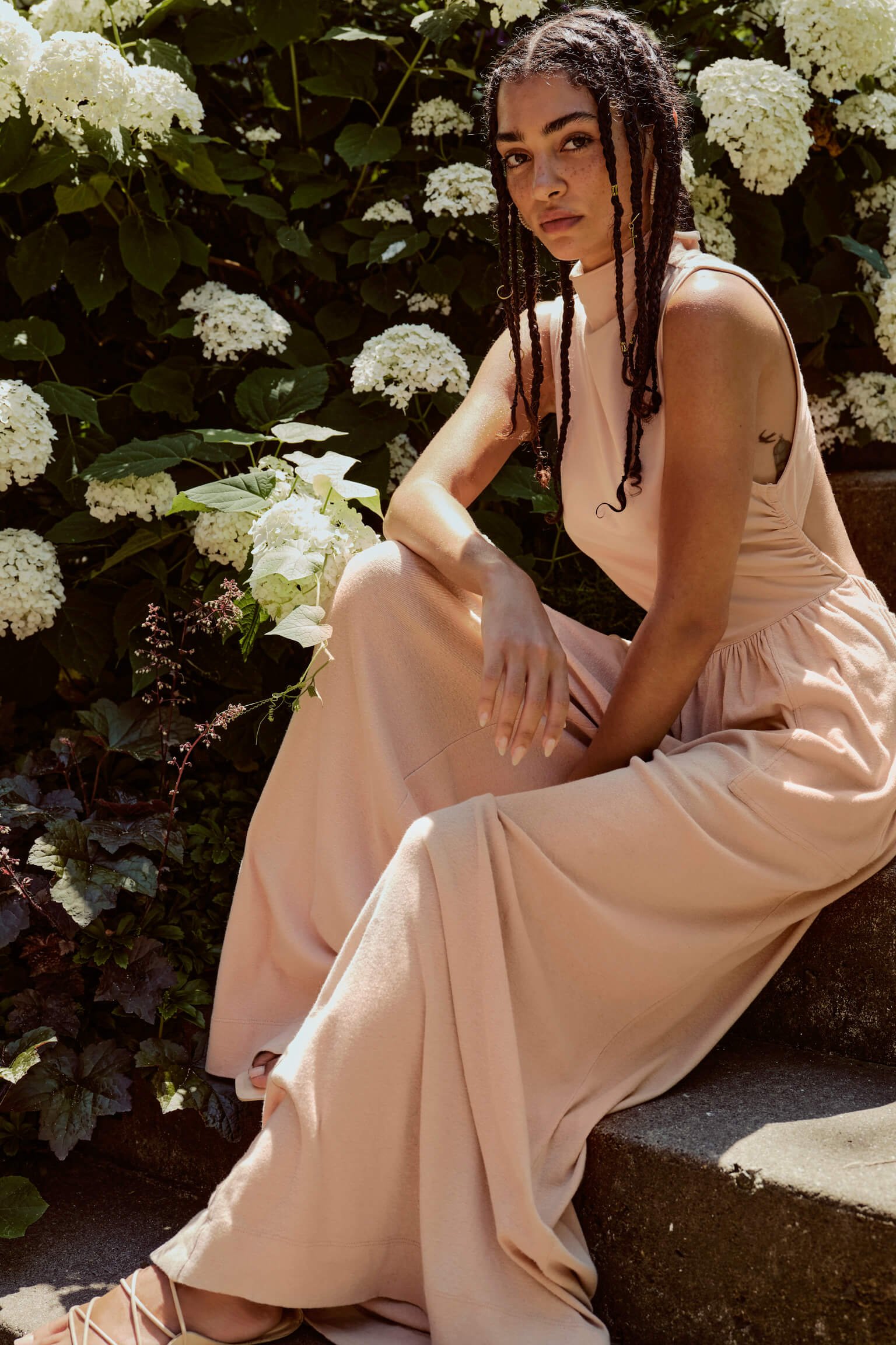 A woman with braided hair in a light pink sleeveless dress sitting on outdoor steps surrounded by white hydrangea flowers.