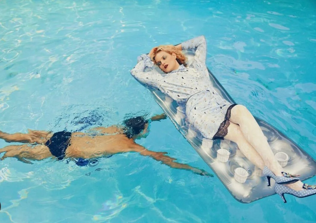 A woman wearing a sleepwear dress lying on an air mattress floating in a swimming pool, with a man swimming nearby underwater.