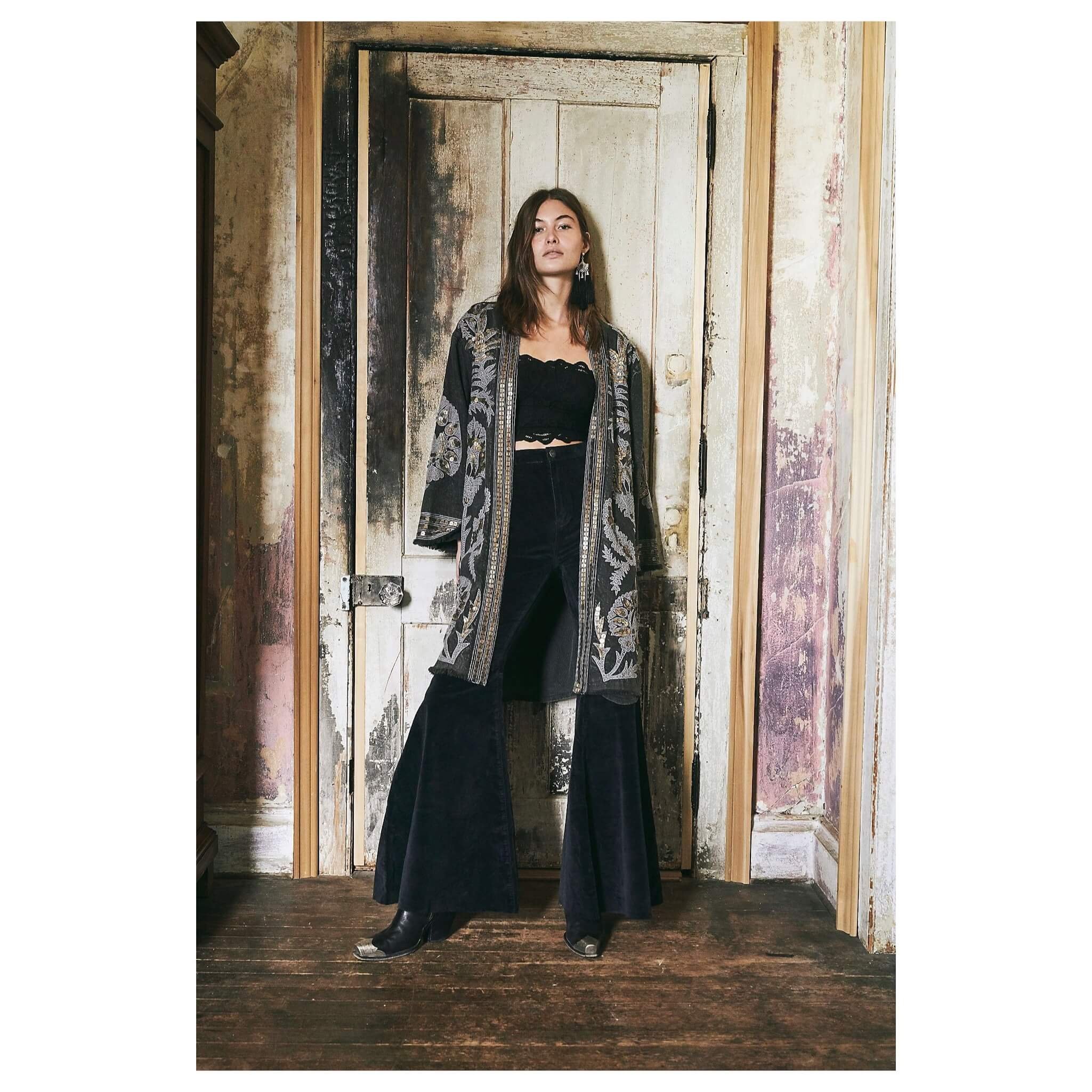 A woman with shoulder-length brown hair standing in front of a rustic wooden door, wearing a black crop top, wide-leg black pants, and an embroidered black coat.