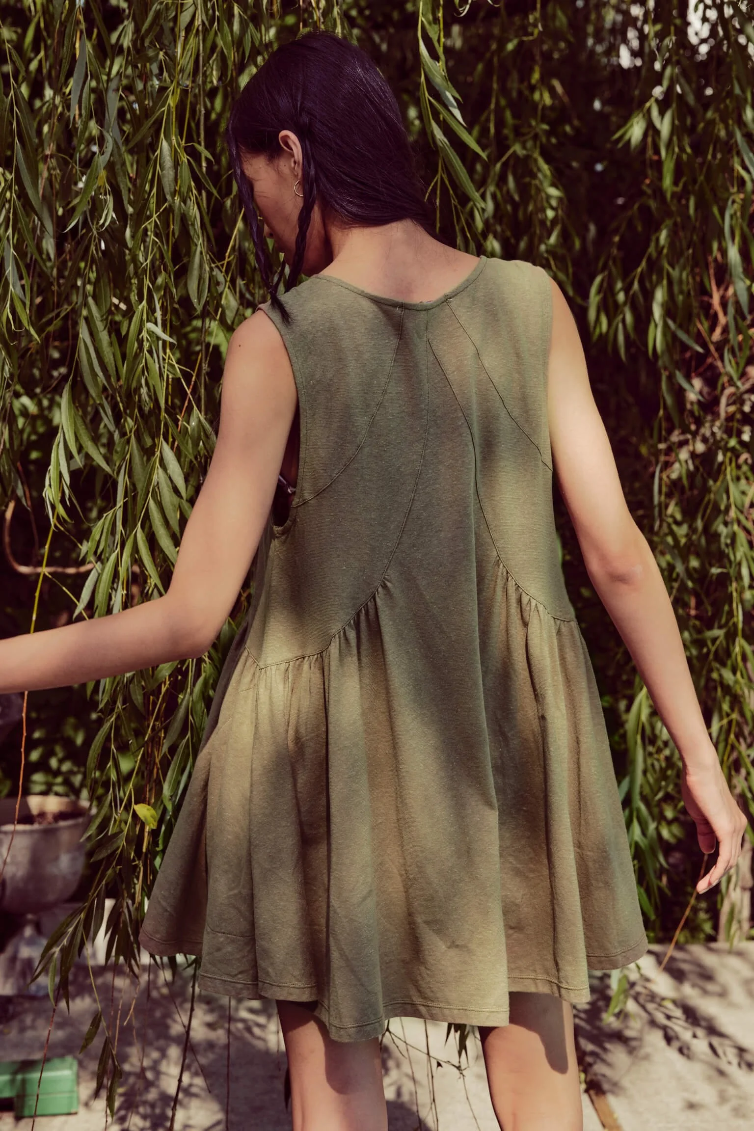 A woman in a sleeveless, olive-green dress standing among hanging branches of a tree, viewed from the back.